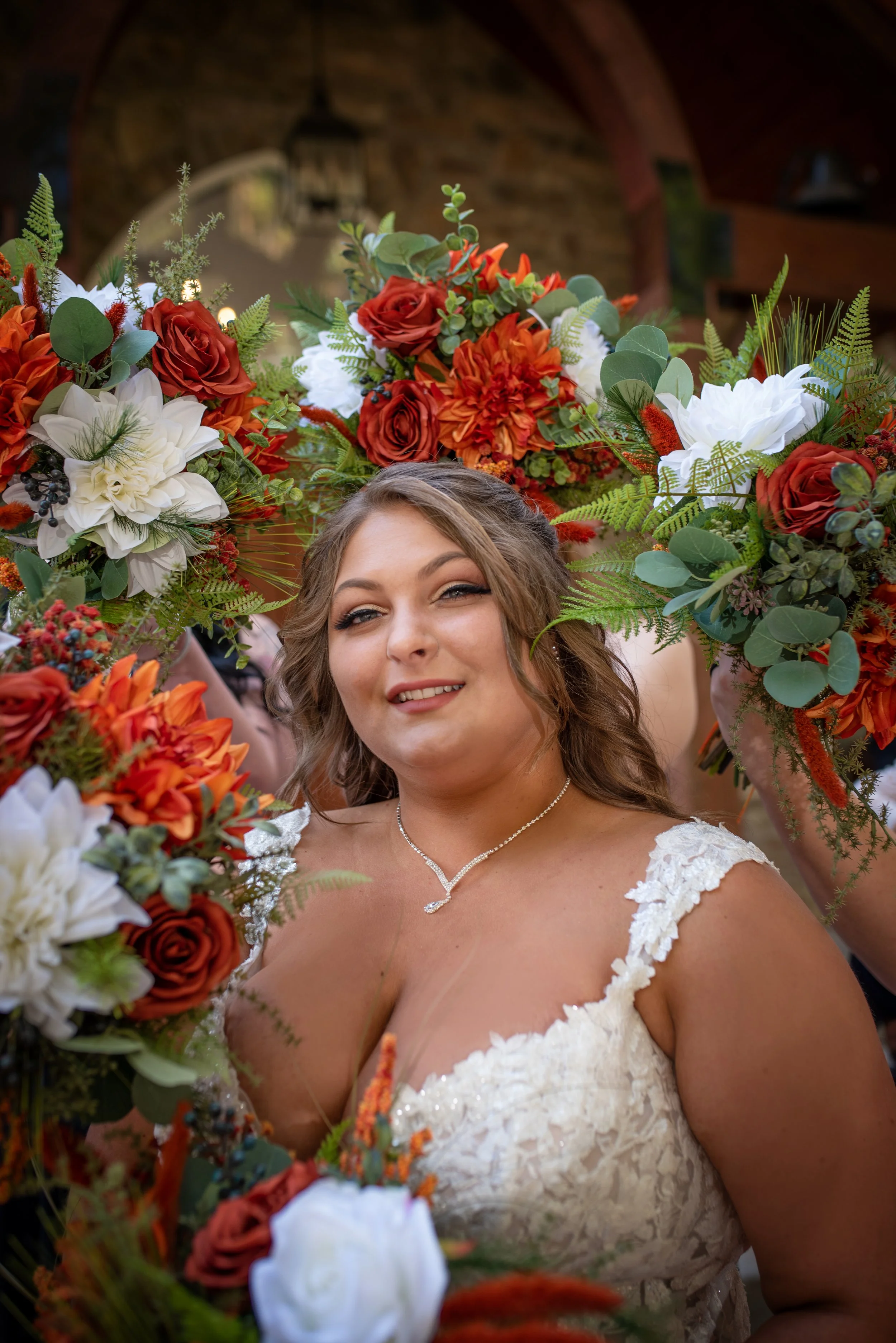 A smiling bride wearing a white lace wedding dress with a sweetheart neckline, standing amidst a colorful floral bouquet with red, white, and orange flowers, greenery, and ferns, in a rustic indoor setting with brick walls and wooden beams.