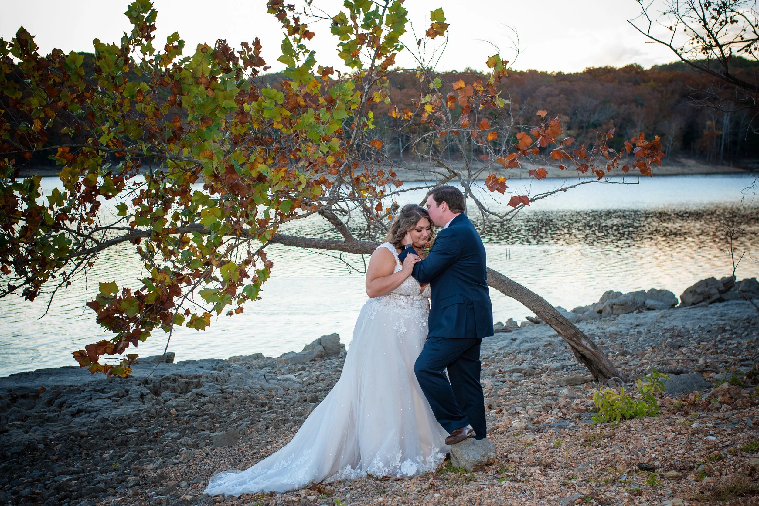 A bride and groom stand by the water's edge, with the groom kissing the bride's forehead. The bride wears a white wedding gown, and the groom wears a dark suit. They are surrounded by autumn-colored trees and rocky terrain near a lake.