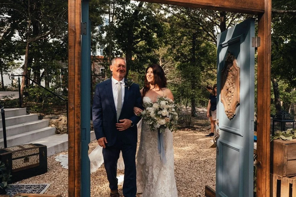 A bride walking down the aisle with an older man, possibly her father, at an outdoor wedding ceremony. The bride holds a bouquet of white flowers, and the setting features trees and decorative doors.