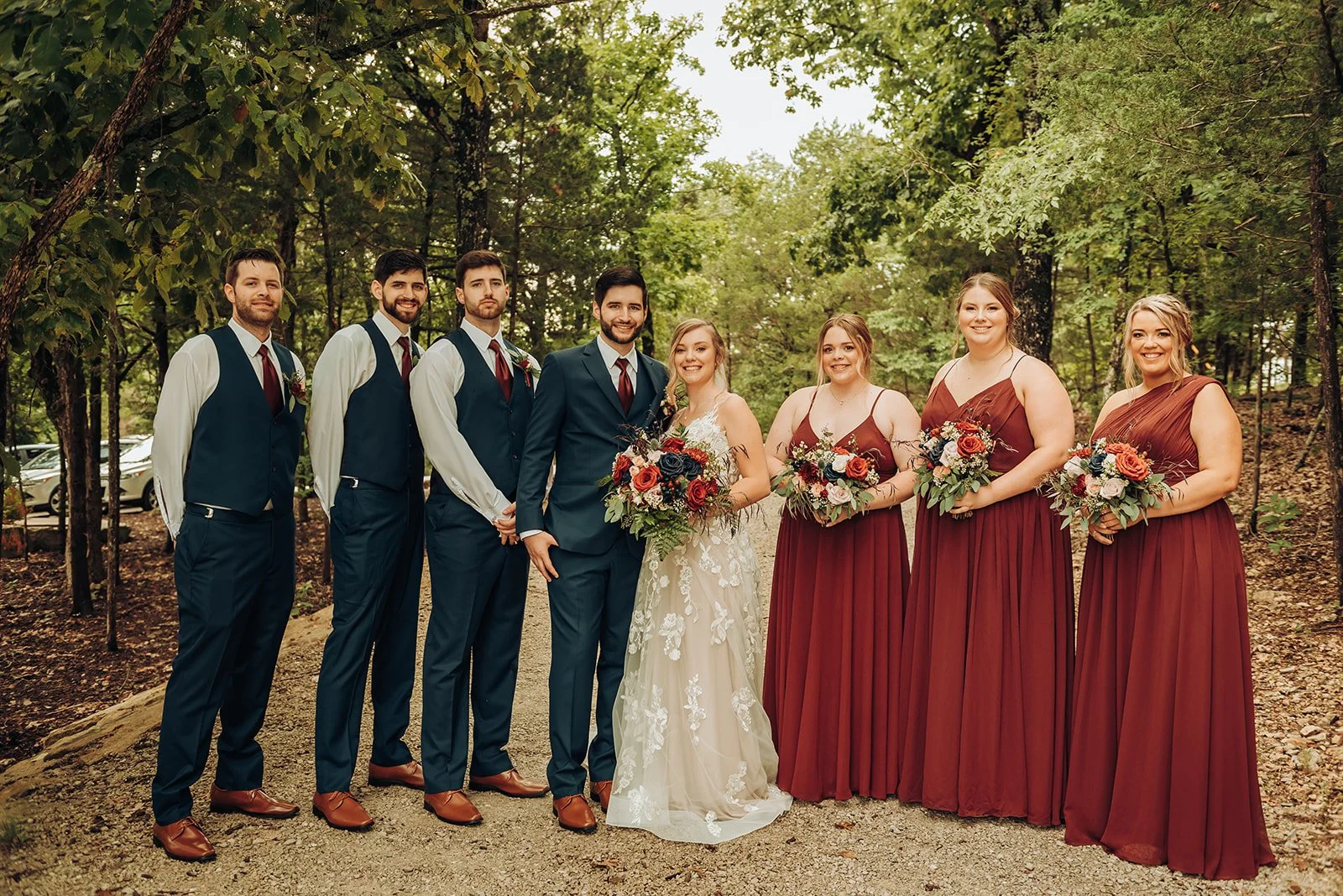 A wedding party standing outdoors on a forest path, consisting of four men in suits and vests, and five women in burgundy dresses, all holding bouquets of flowers.