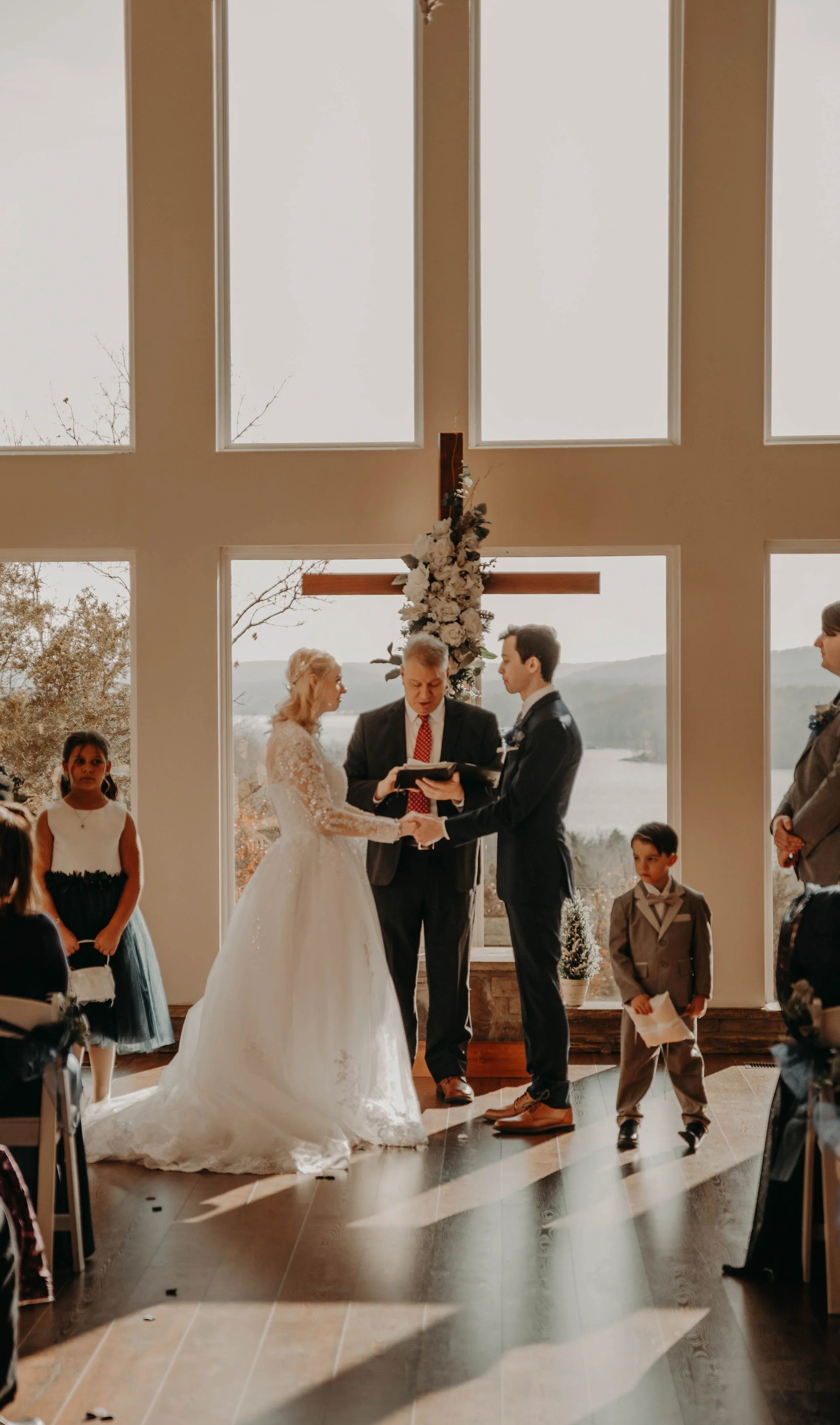 A wedding ceremony taking place indoors with large windows and an outdoor view. The bride and groom are exchanging vows, holding hands, standing in front of a floral cross backdrop. Guests and ring bearers are present, and sunlight streams through the windows, illuminating the scene.