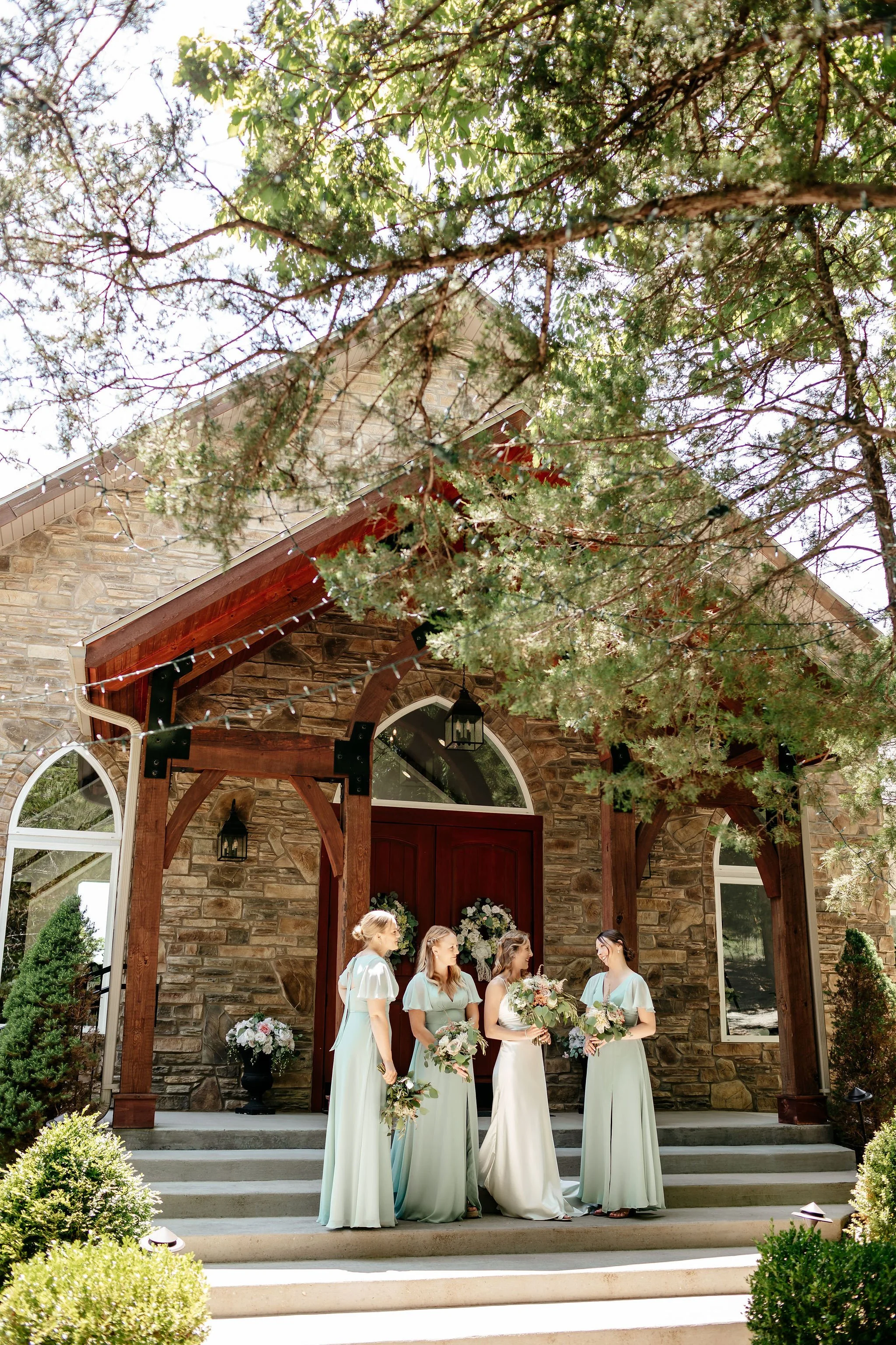 Four women in light blue dresses standing on the steps of a stone house with a wooden porch, holding bouquets of flowers, under a tree decorated with string lights, during an outdoor wedding ceremony.