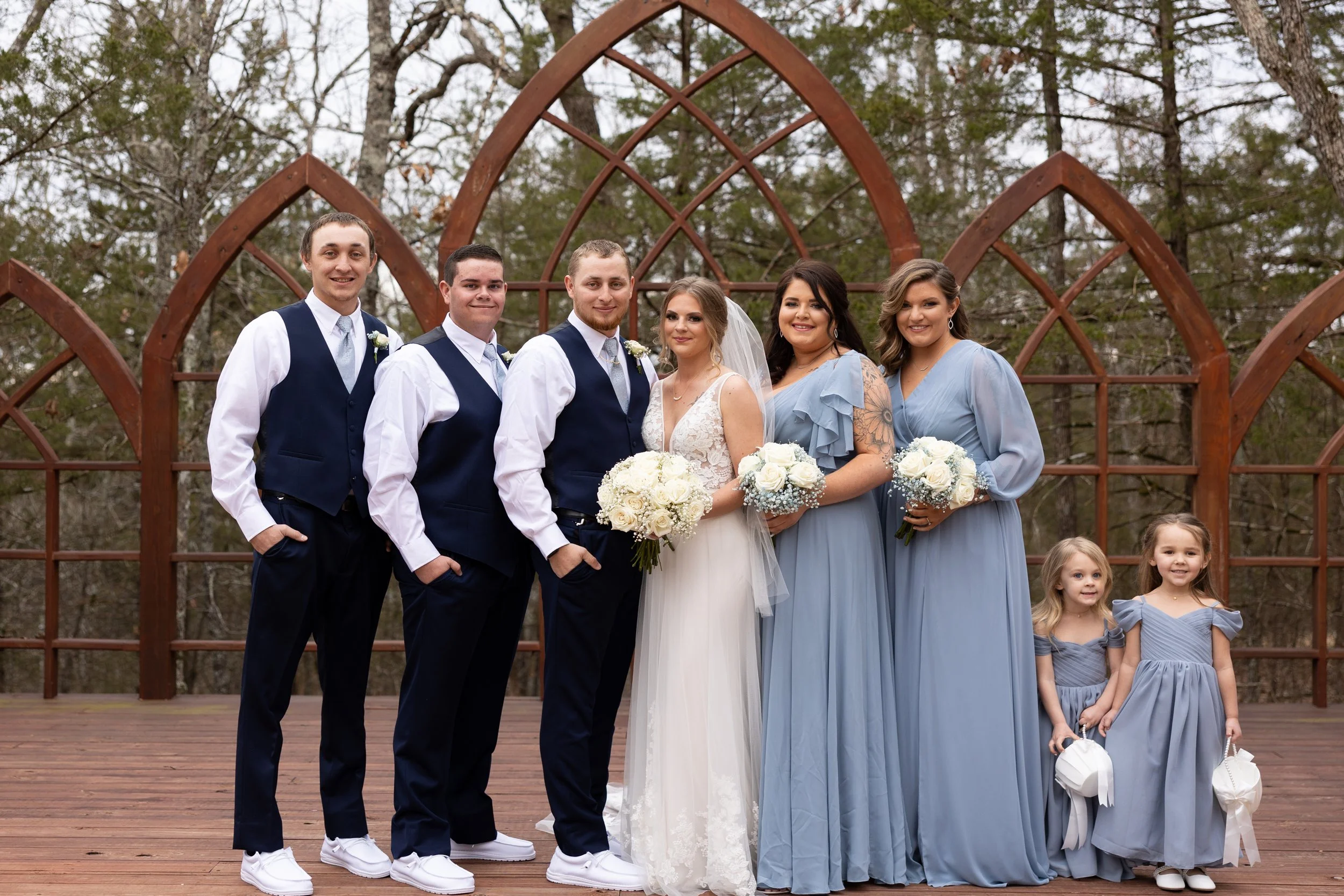 A wedding group photo outdoors with six adults and three children in front of a decorative wooden structure and trees.