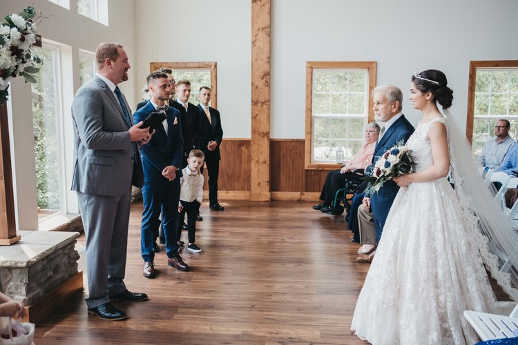 Bride in a white wedding gown holding a bouquet standing opposite a group of men in suits, with elderly seated guests in a wooden-paneled room with large windows.