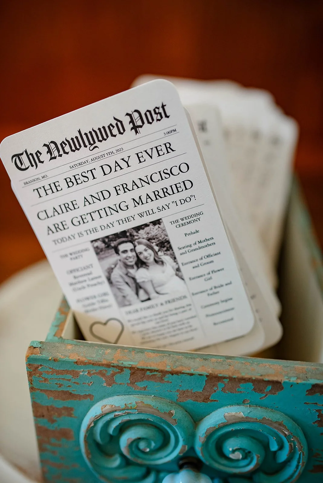 A newspaper-style wedding program titled 'The Newlgwed Post' with a headline 'The Best Day Ever' about Claire and Francisco getting married, including a black-and-white photo of the couple and details on the wedding ceremony schedule, resting in a de