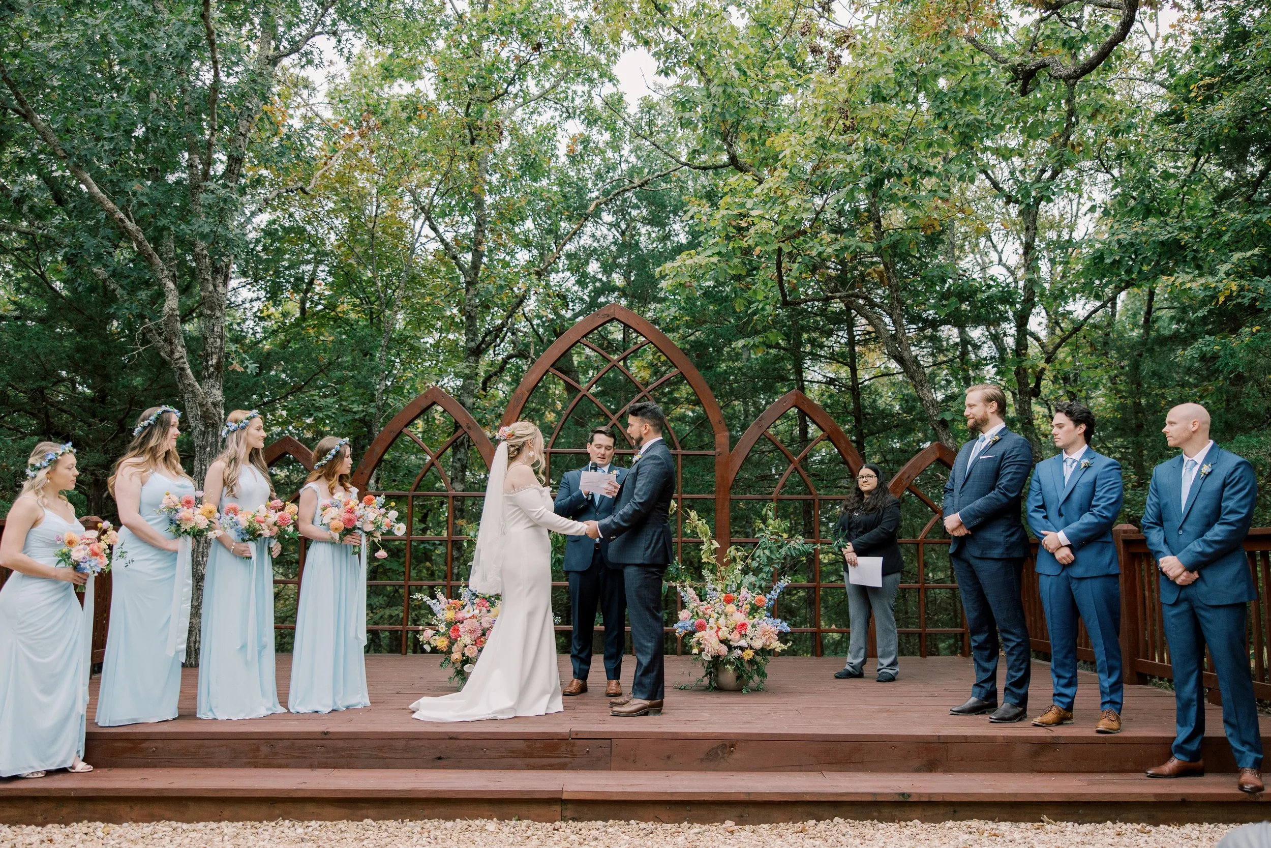A wedding ceremony taking place outdoors in a wooded area, with a bride and groom exchanging vows in front of an officiant, surrounded by bridesmaids in light blue dresses holding bouquets, and groomsmen in blue suits standing nearby.