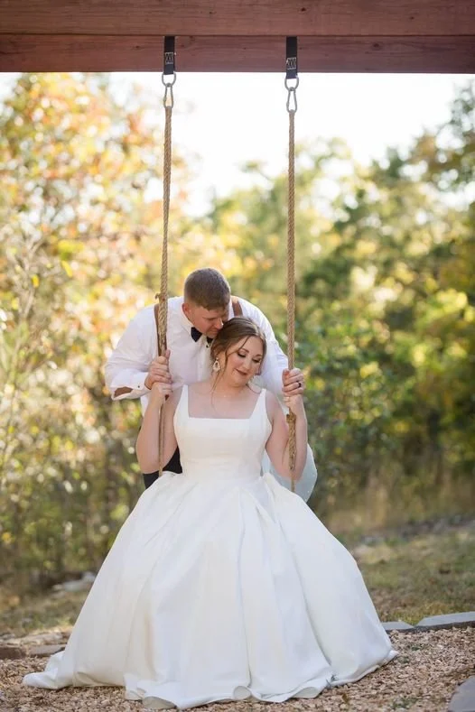 A bride in a white wedding dress sitting on a swing, with a groom in a white shirt and black bow tie standing behind her, holding the swing's ropes outdoors during daytime.