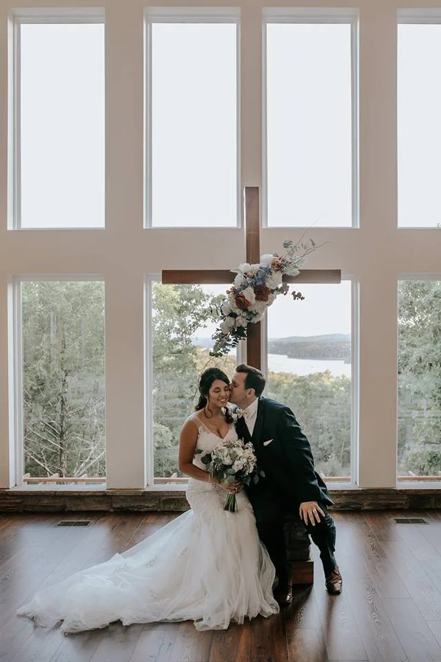 A bride and groom in wedding attire in front of a large cross decorated with flowers, inside a building with large windows overlooking a natural landscape.