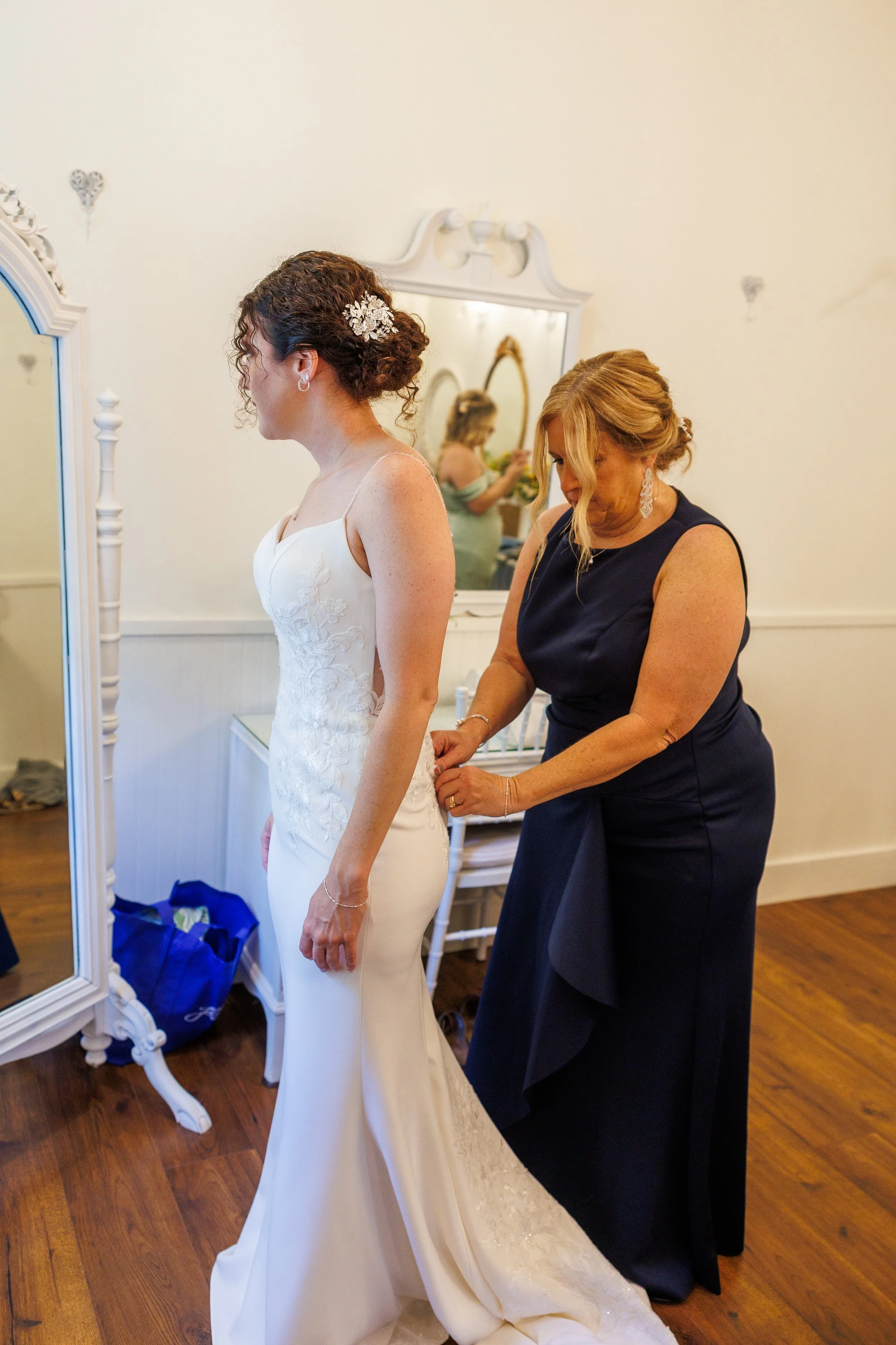 A bride in a white wedding dress standing in front of a mirror, with a woman in a navy blue dress fastening the back of her dress.