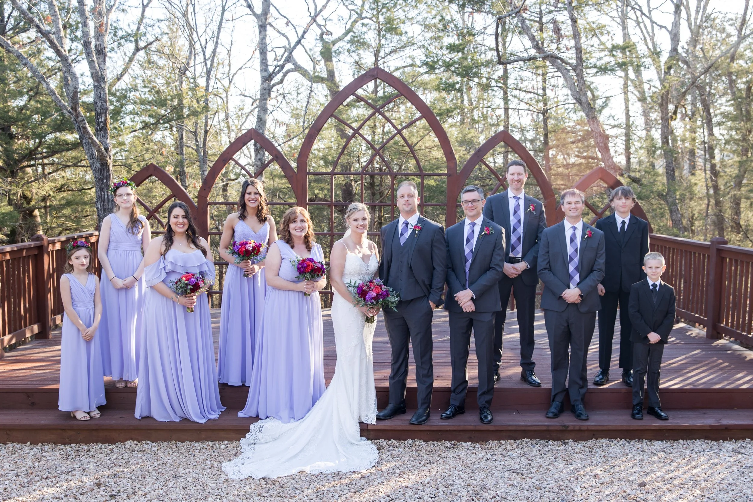 A group of people posing on a wooden deck surrounded by trees, with a wedding theme. The bride is in a white wedding dress, holding a bouquet, and the groom is in a dark suit. Bridesmaids are in lavender dresses and holding bouquets, and groomsmen are in dark suits with purple accents.
