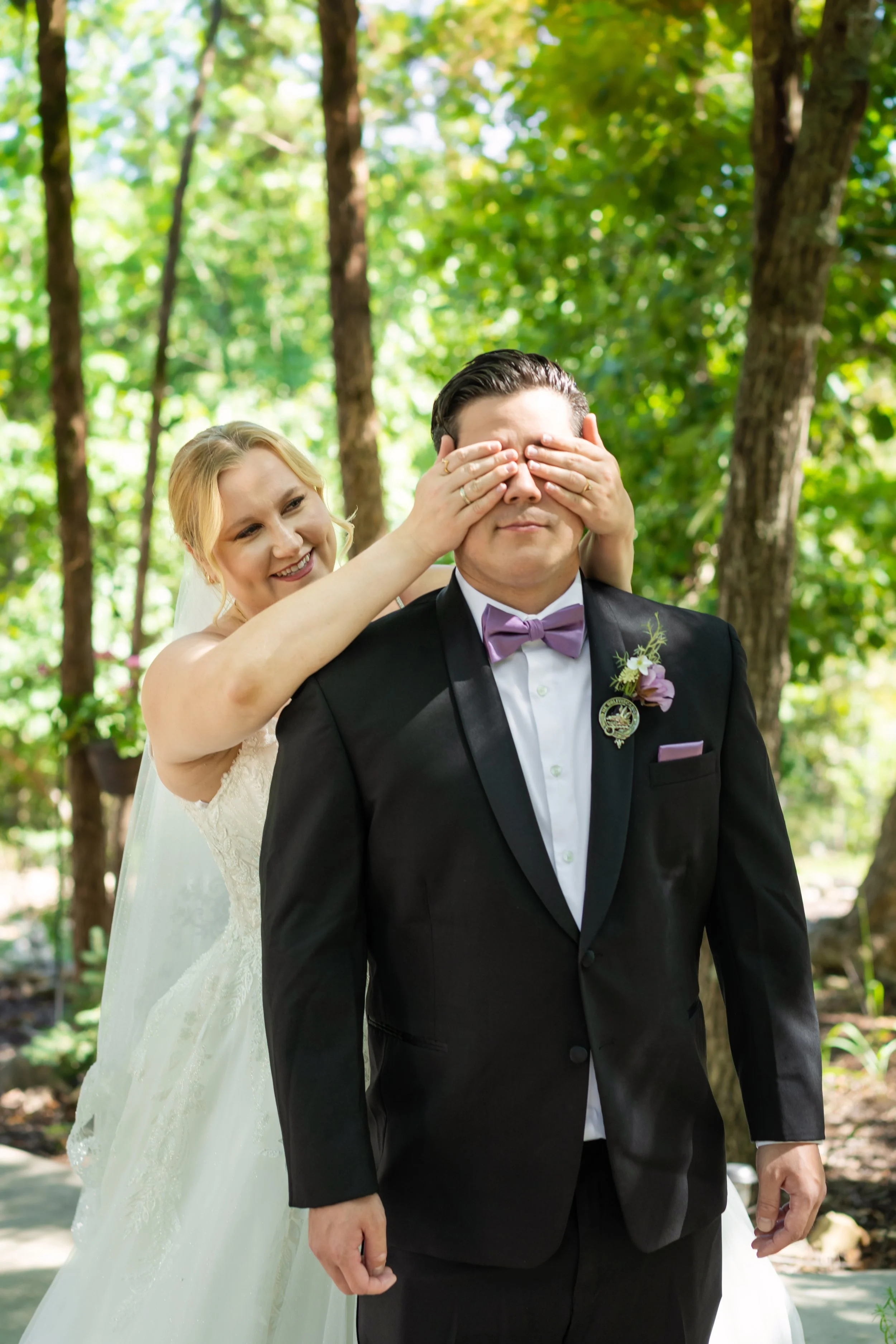 A bride with blonde hair and a white wedding dress smiles as she playfully covers a groom's eyes from behind, who is dressed in a black tuxedo with a purple bow tie, in an outdoor setting with green trees.