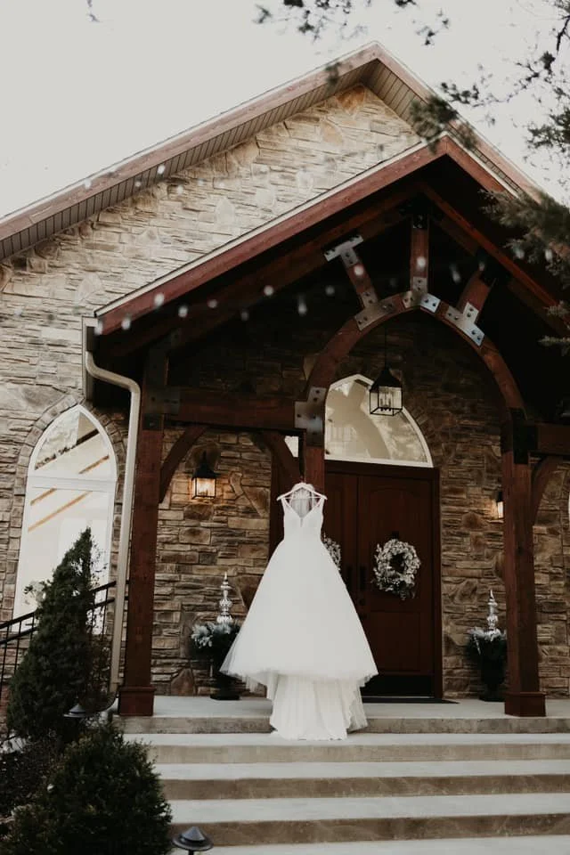 A white wedding dress hanging in front of a rustic brick house decorated for the holidays, with wreath on the door and potted plants on the steps.