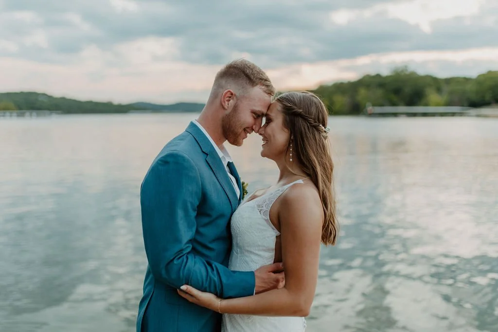 A couple dressed in wedding attire standing close together by a large body of water with trees and a cloudy sky in the background.