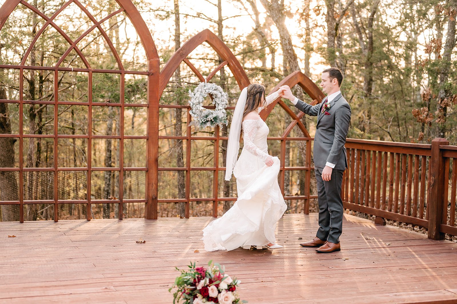 A bride and groom dancing on an outdoor wooden deck during their wedding ceremony, with trees and a decorative wooden arch in the background, and a bouquet of flowers on the ground in the foreground.