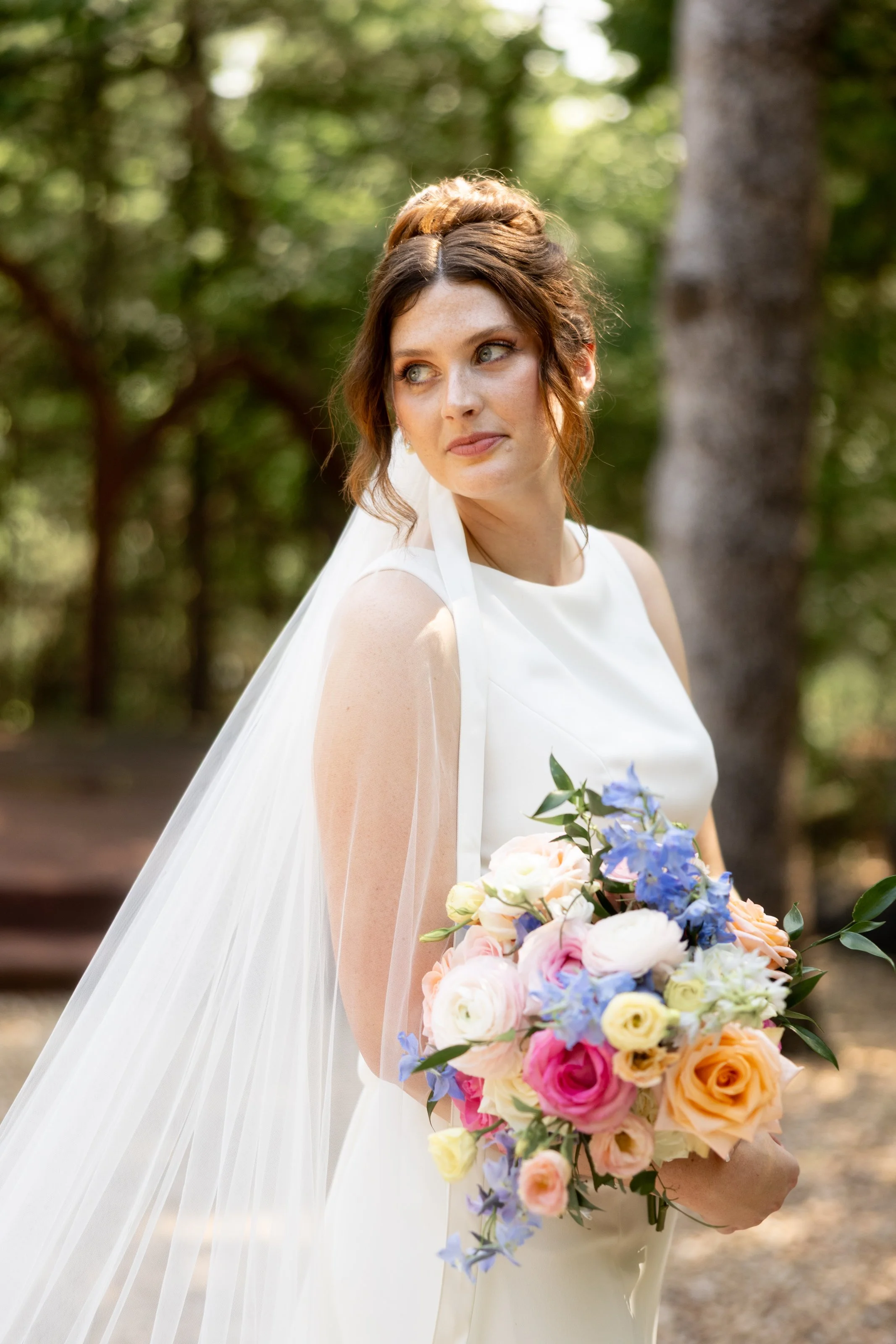 A woman in a white dress holding a bouquet of colorful flowers outdoors surrounded by trees.