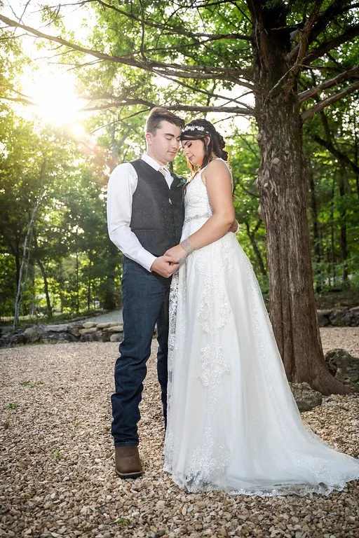 A bride and groom standing outdoors under a large tree, holding hands with their foreheads touching, during a wedding photo shot in a wooded area with sunlight filtering through the leaves.