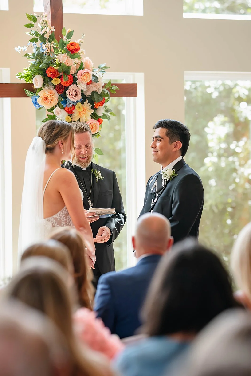 A bride and groom exchanging vows during a wedding ceremony, with a officiant reading from a book, in a room decorated with a large floral arrangement, and guests seated in front of them.