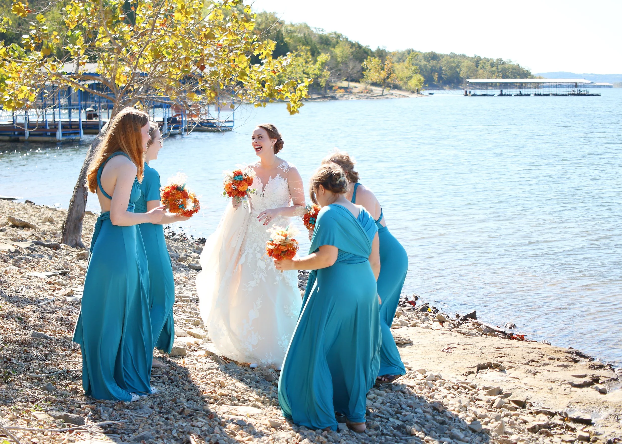 A bride in a white wedding dress and bridesmaids in turquoise dresses standing on a rocky shore by a lake, smiling and holding bouquets, with trees and a dock in the background.