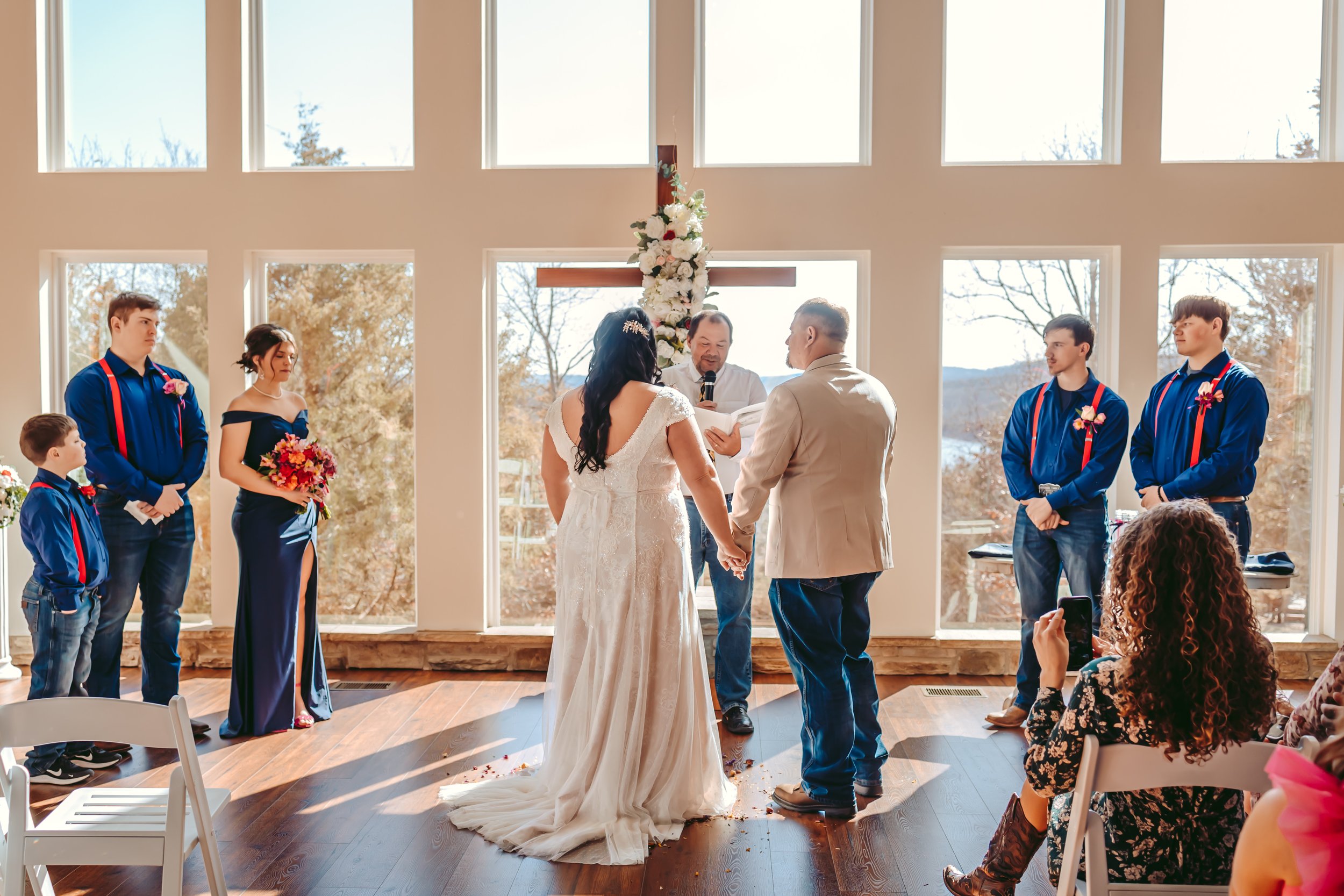 A wedding ceremony taking place indoors near large windows with trees outside. The bride and groom are holding hands, facing each other, with an officiant in between them. The wedding party members are standing on either side, dressed in dark blue ou