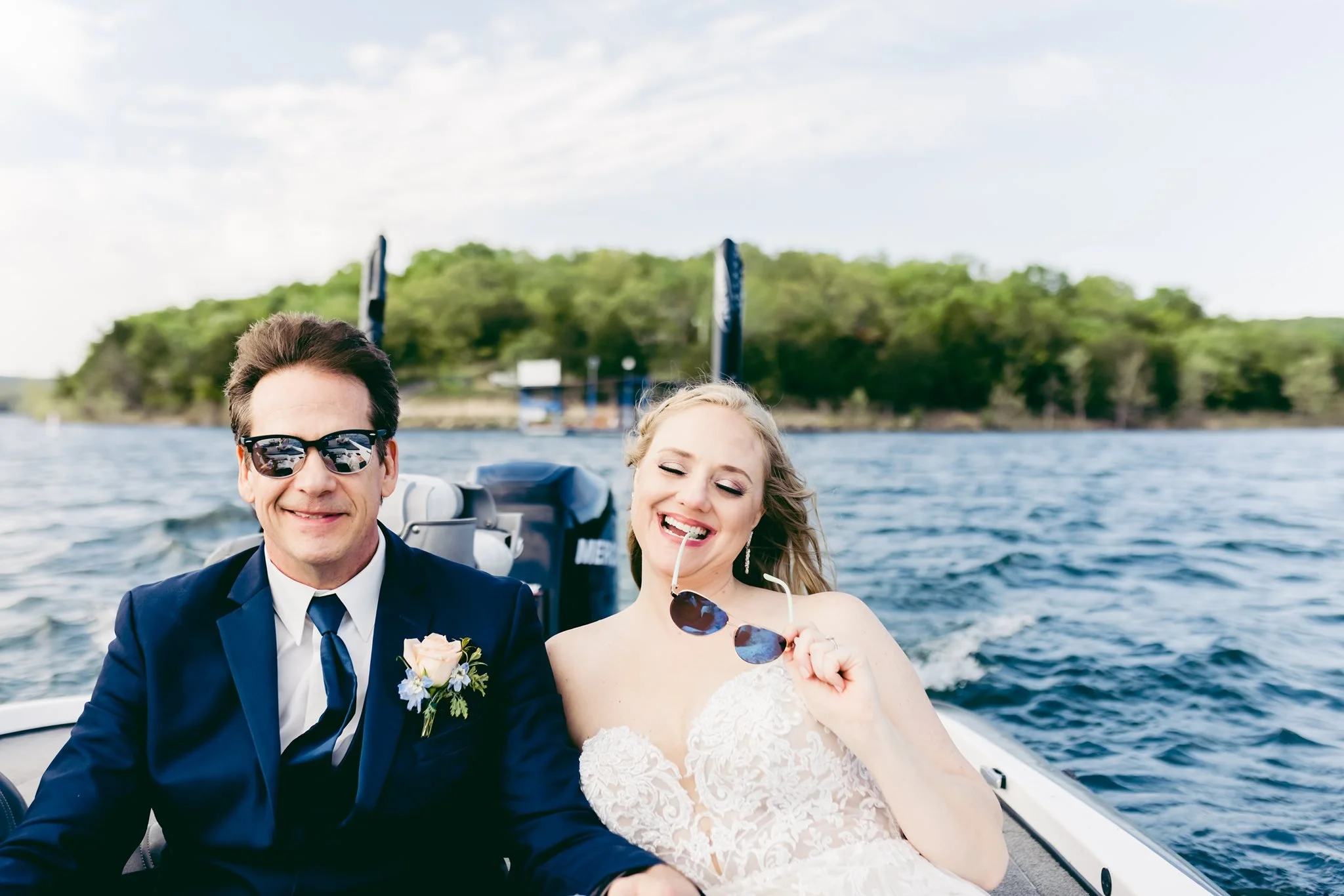 A joyful couple in wedding attire enjoying a boat ride on the water, with a lush green island and a partly cloudy sky in the background.