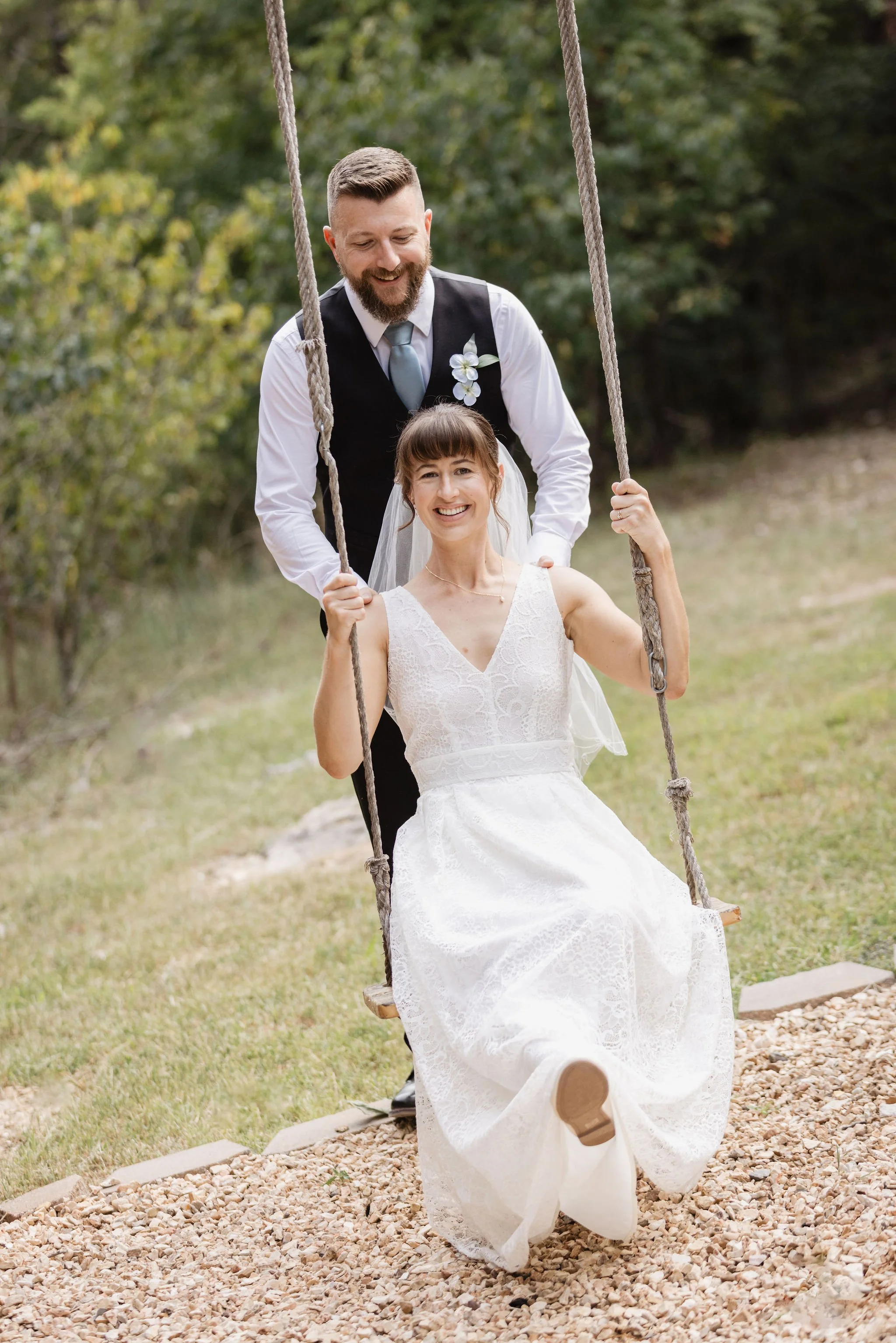 A man and woman on a swing set outdoors with trees in the background, the woman wearing a white lace wedding dress and the man in a suit with a boutonniere, both smiling.