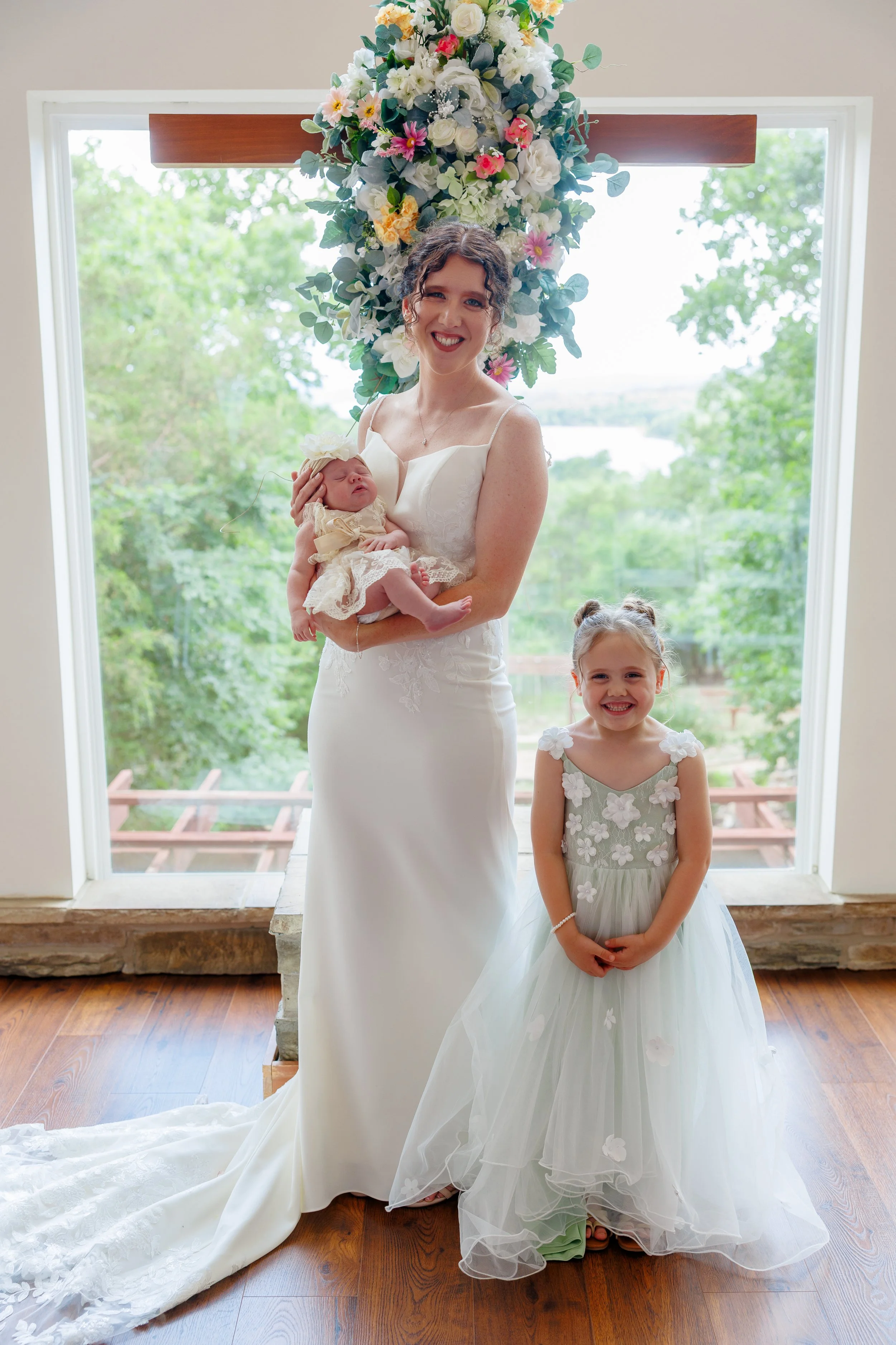 A woman in a wedding dress holding a newborn baby, standing next to a young girl in a dress with floral details, in front of a window with a floral cross decoration overhead.