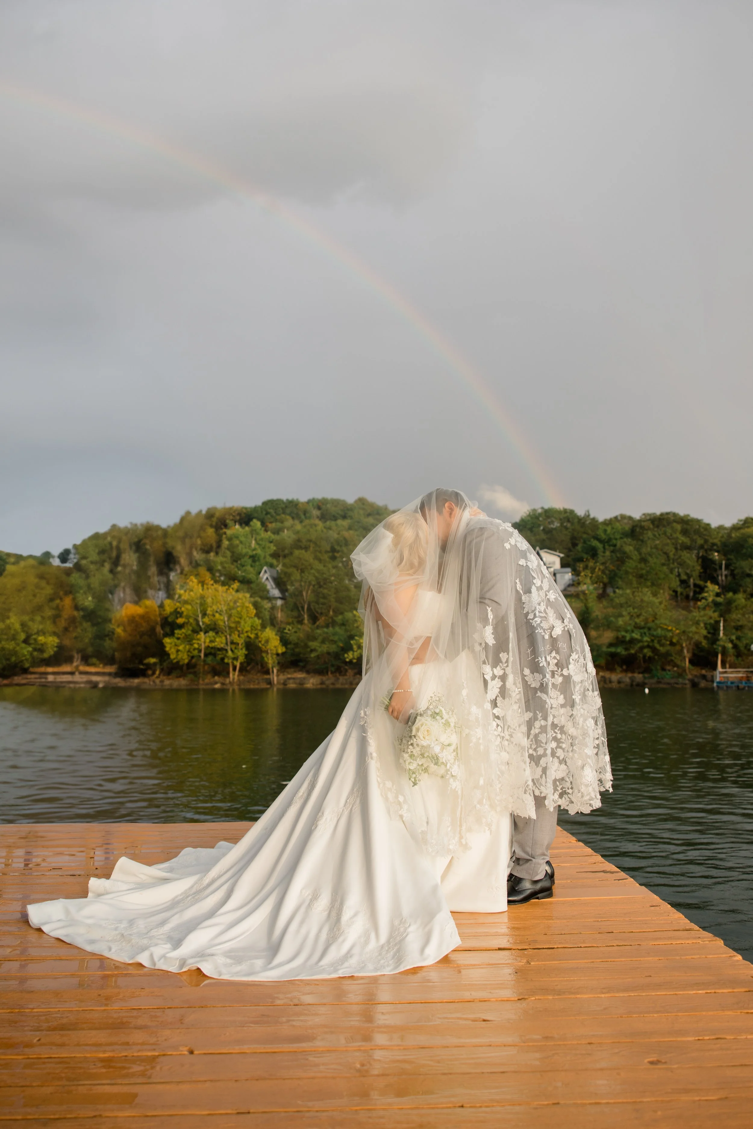 A bride and groom share a kiss on a wooden dock by a lake, with a colorful rainbow and cloudy sky in the background.