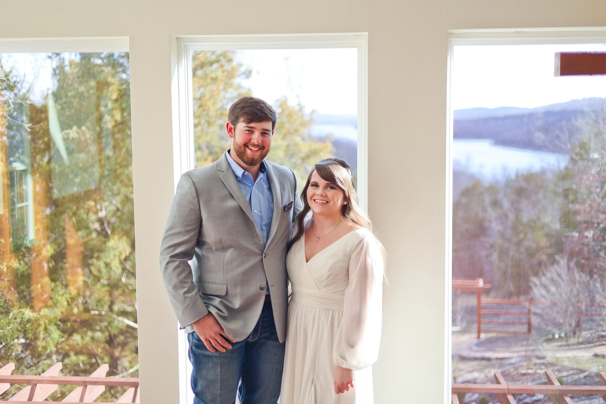 A smiling young couple standing inside near large windows overlooking a river and trees. The man is wearing a light grey blazer and blue shirt, and the woman is in a white dress.