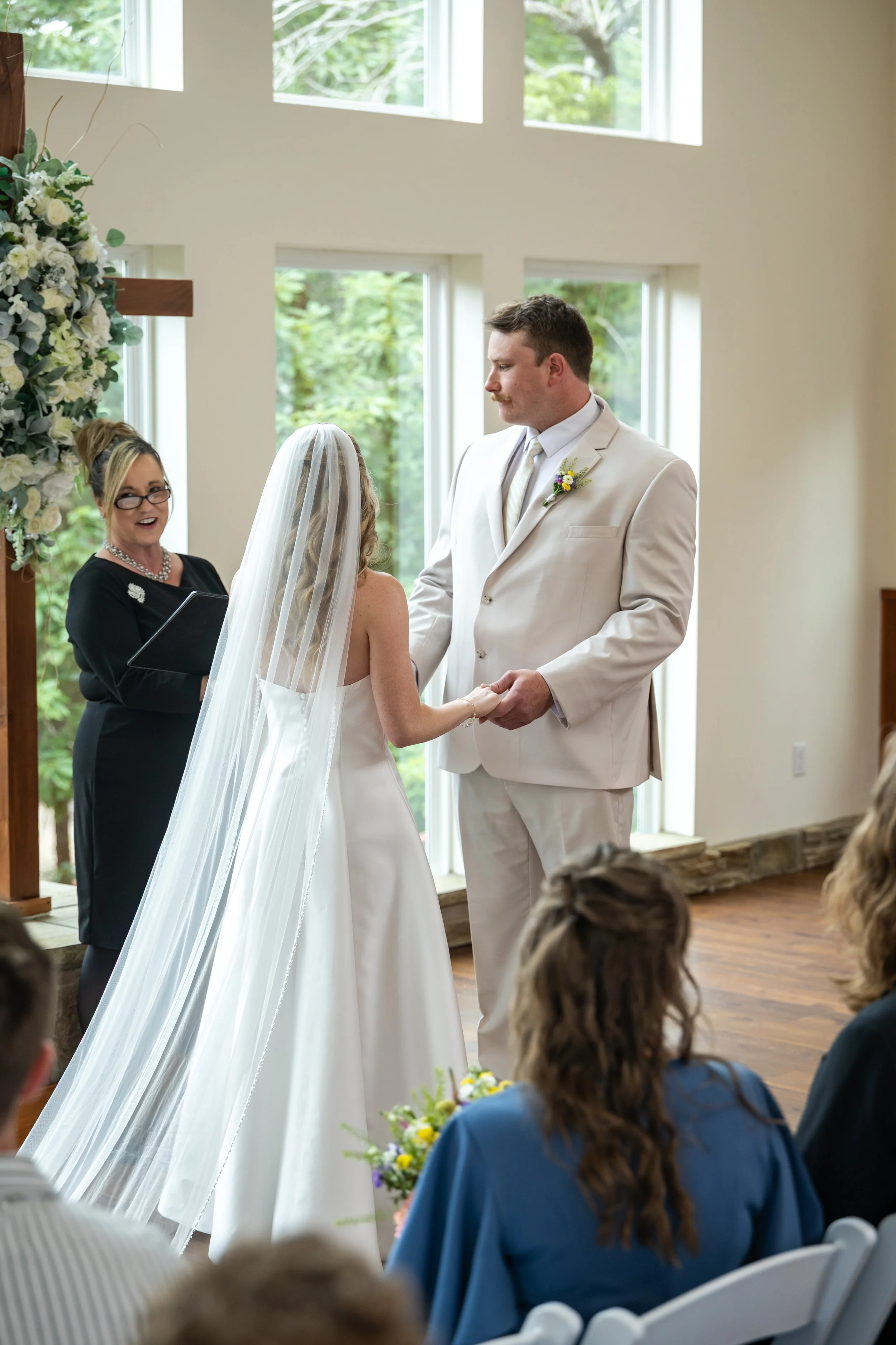 A wedding ceremony with a bride and groom holding hands and facing each other in front of an officiant, inside a room with large windows and greenery outside.
