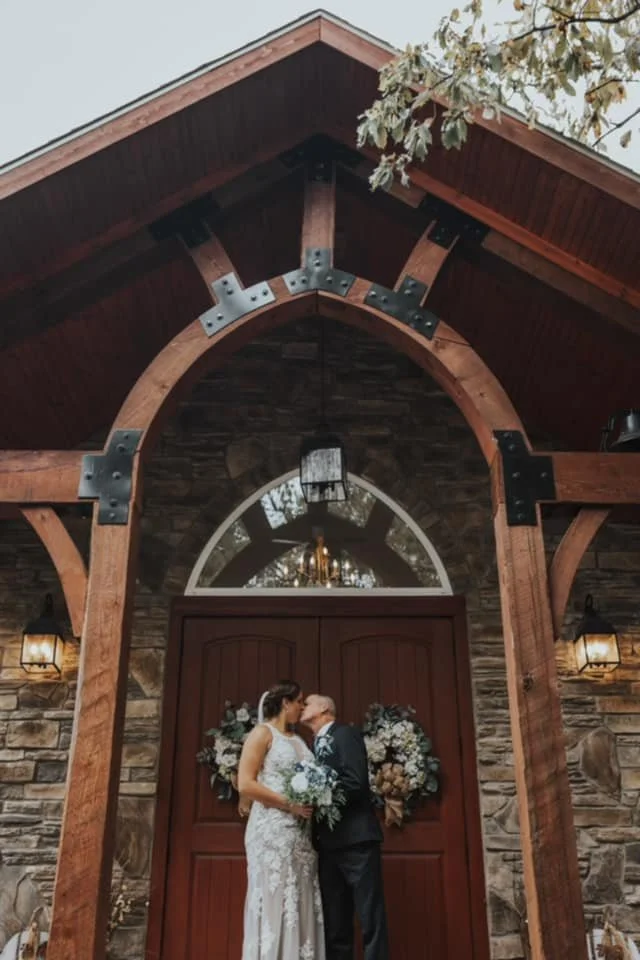 A bride and an older man, likely her father, sharing a kiss in front of a large wooden door at a rustic wedding venue with stone walls and decorative wreaths.
