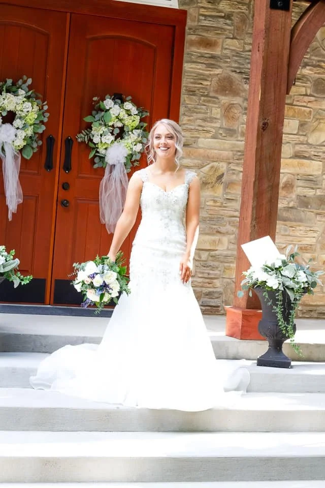 Bride in a white wedding dress holding a bouquet, standing on the steps outside a building with wooden doors decorated with floral arrangements.