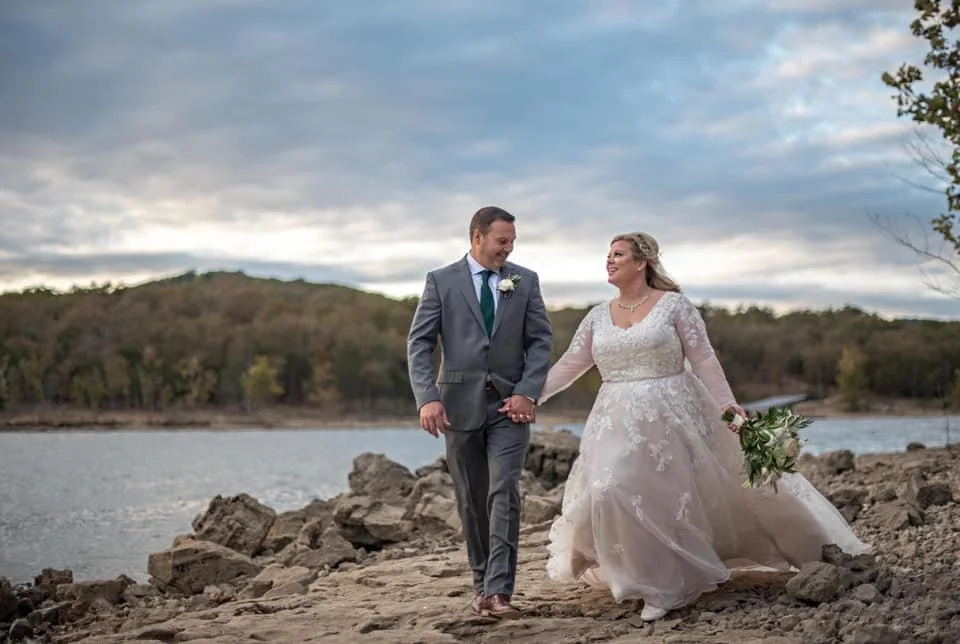 A newlywed couple walking hand in hand along a rocky lakeside, with trees and cloudy sky in the background.