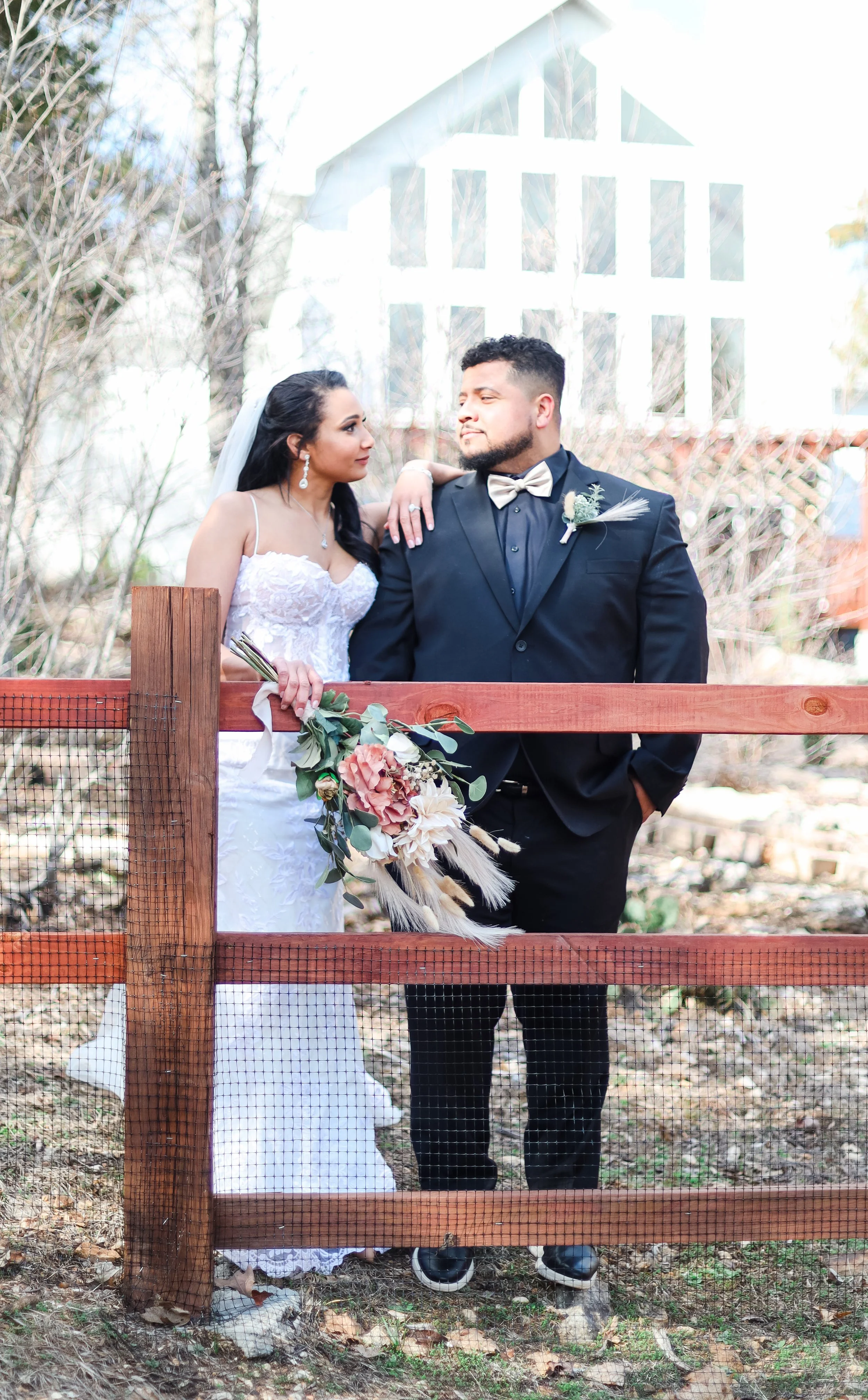 A bride and groom in wedding attire standing behind a wooden fence with a mesh screen, outdoors with trees and a building in the background.