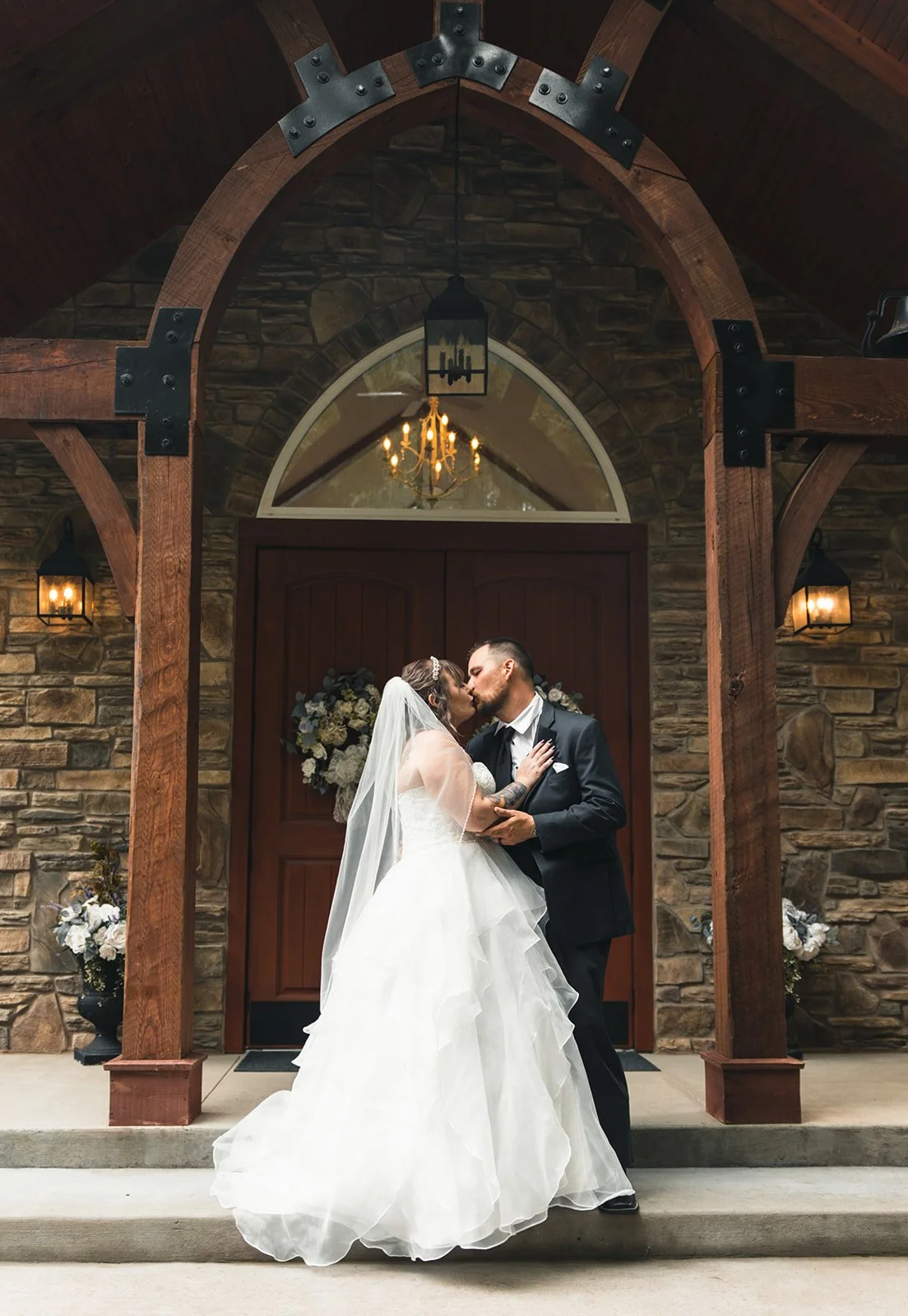 A bride and groom sharing a kiss on the steps of a church or wedding venue, with a stone facade, wooden doors, floral arrangements, and hanging light fixtures.