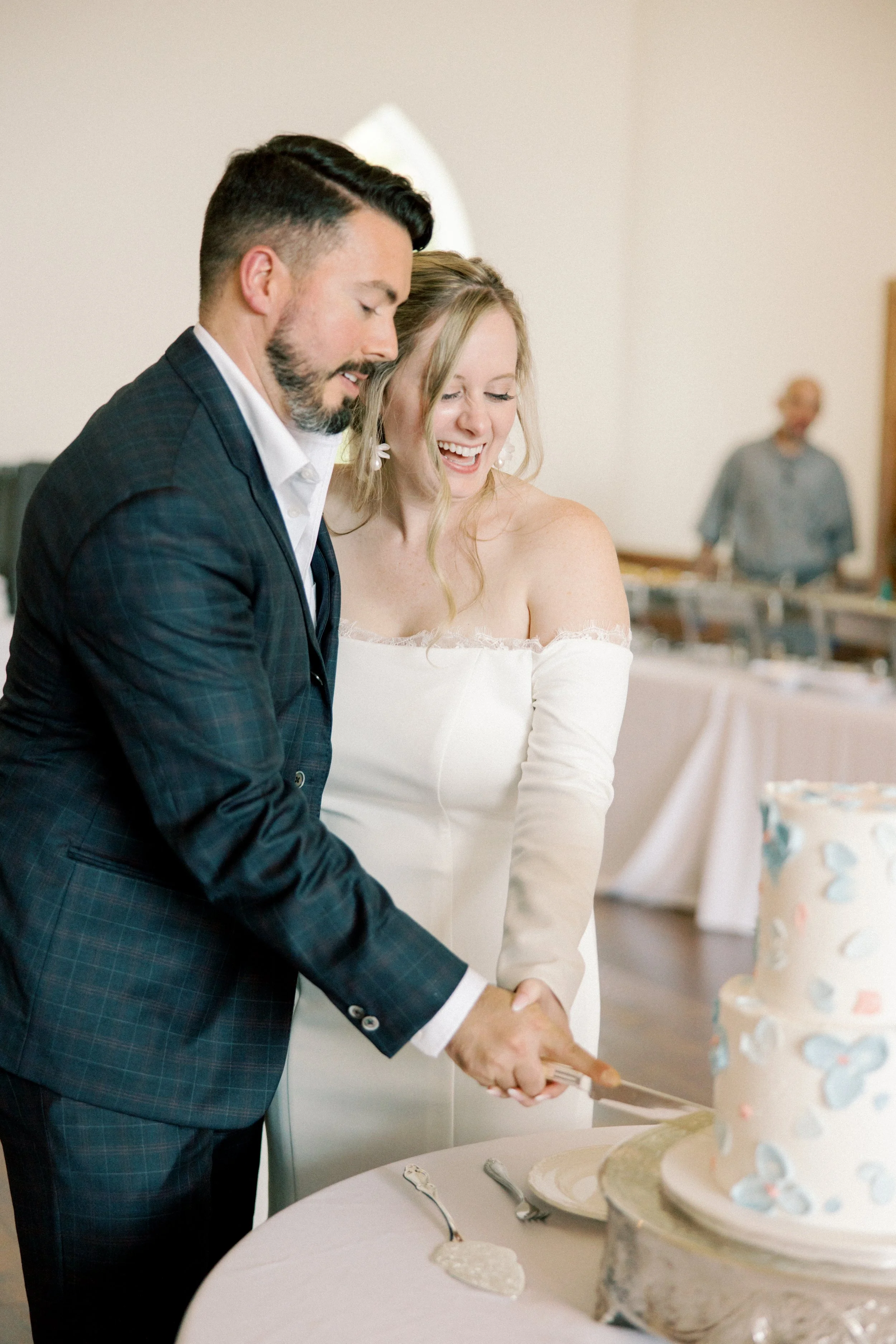 A man and woman in wedding attire cutting a wedding cake together at a celebration, with a person in the background.
