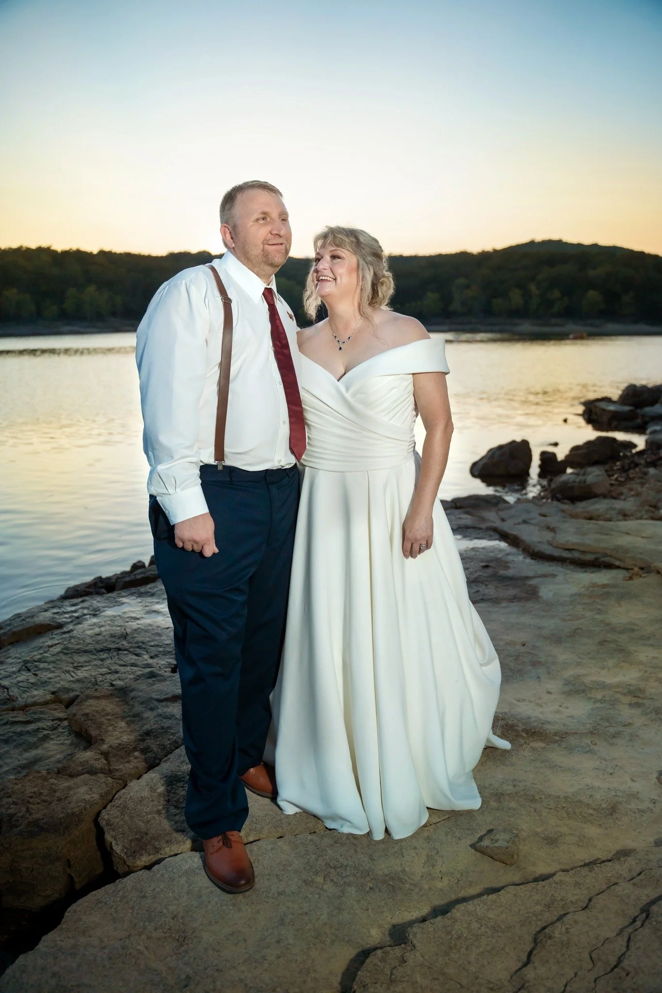 A bride and groom stand on rocks near a body of water during sunset, smiling and looking at each other. The bride wears an off-the-shoulder white wedding dress, and the groom wears a white shirt, red tie, and suspenders.
