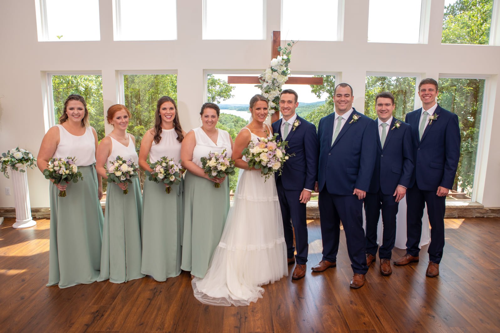 A wedding party inside a building with large windows showing trees and a lake in the background. The group includes the bride in a white dress holding a bouquet, groom in a navy suit, four bridesmaids in light green skirts and white tops, and three g