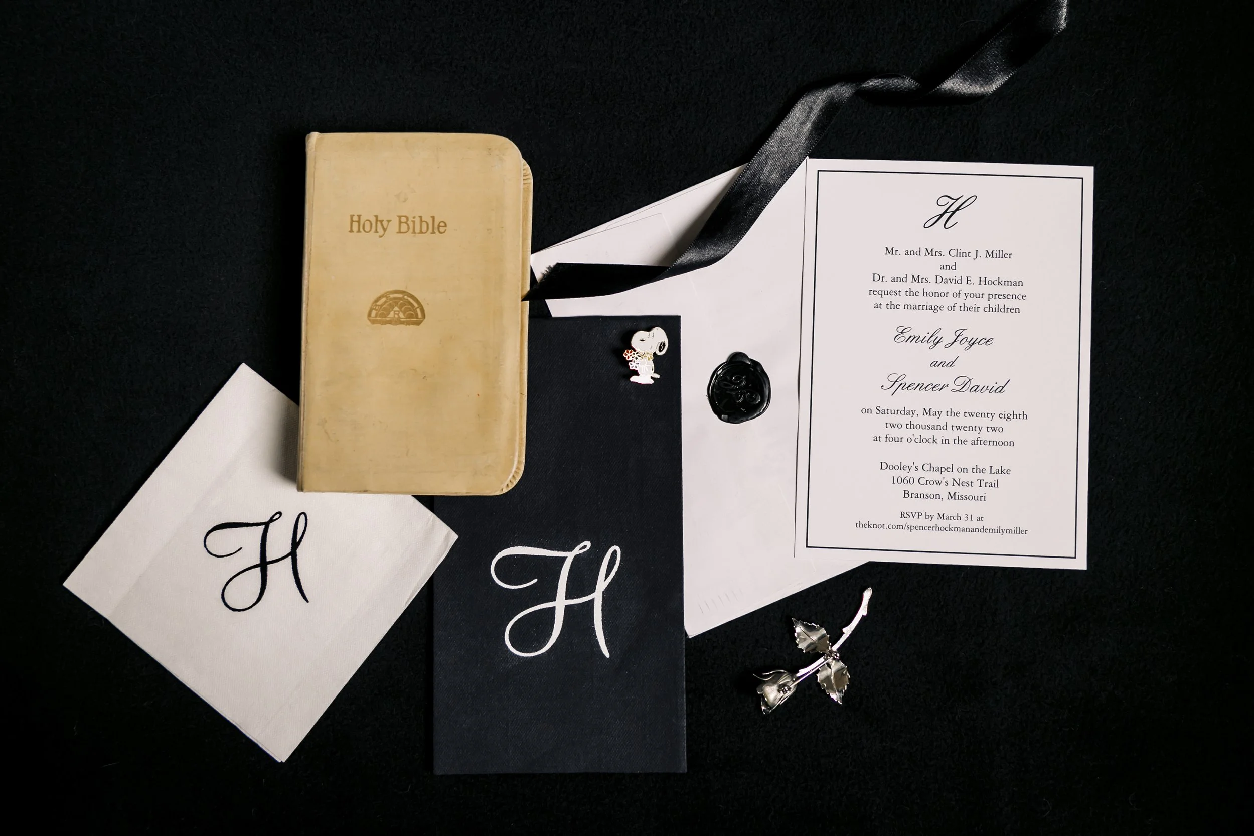Wedding invitation, Holy Bible, peacock feather earrings, and wedding details on black background.