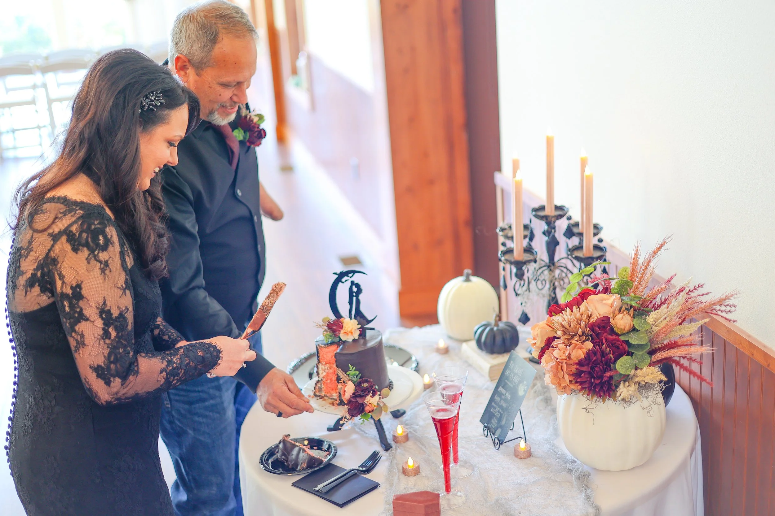 A young woman and an older man are standing together, looking down at a table with desserts and candles during a celebration. The woman wears a black lace dress with a hair accessory, and the man wears a dark shirt with a boutonniere. The table has a