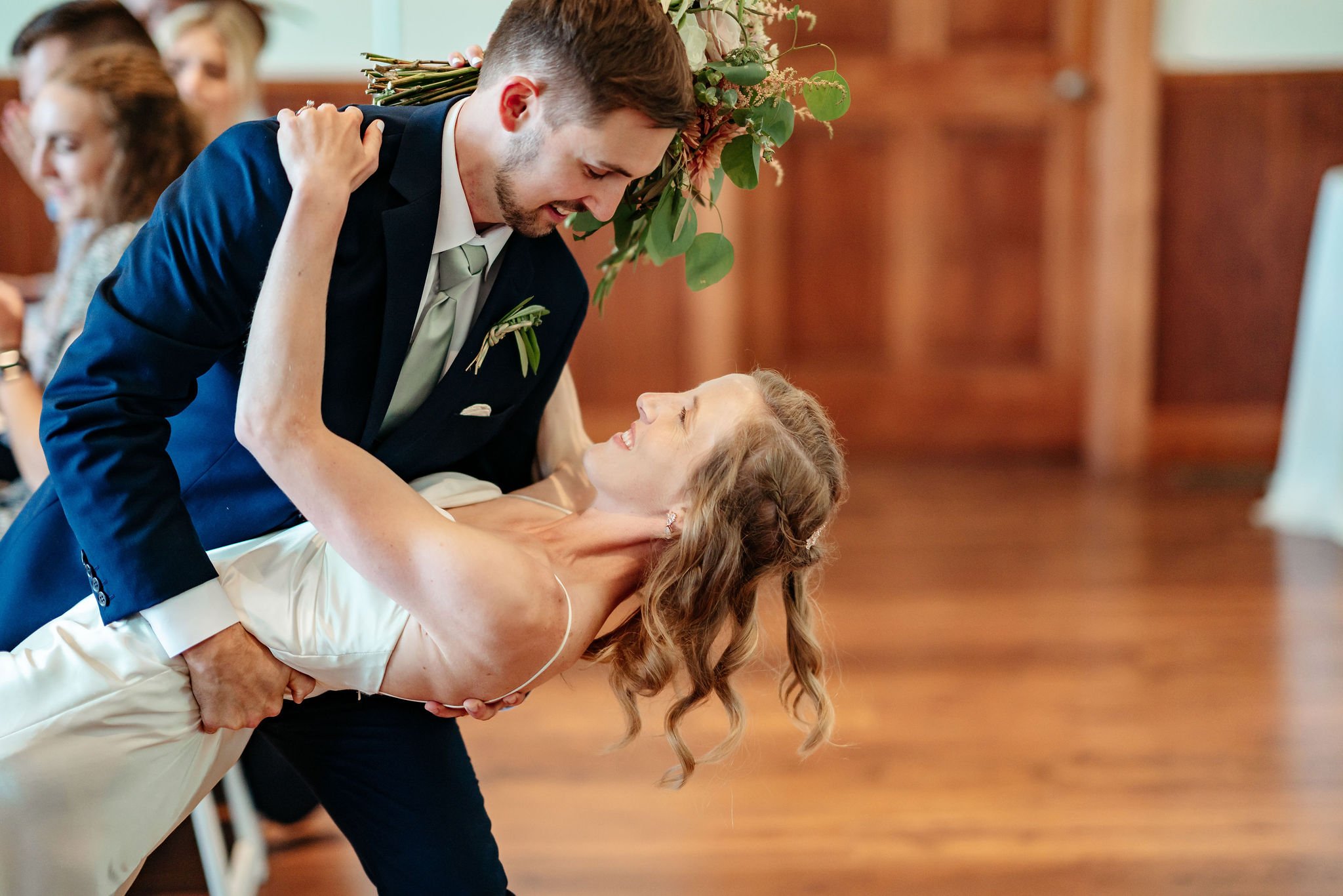 A man in a suit and a woman in a white dress are dancing together at a wedding reception, with the man holding the woman in a dip and both smiling.