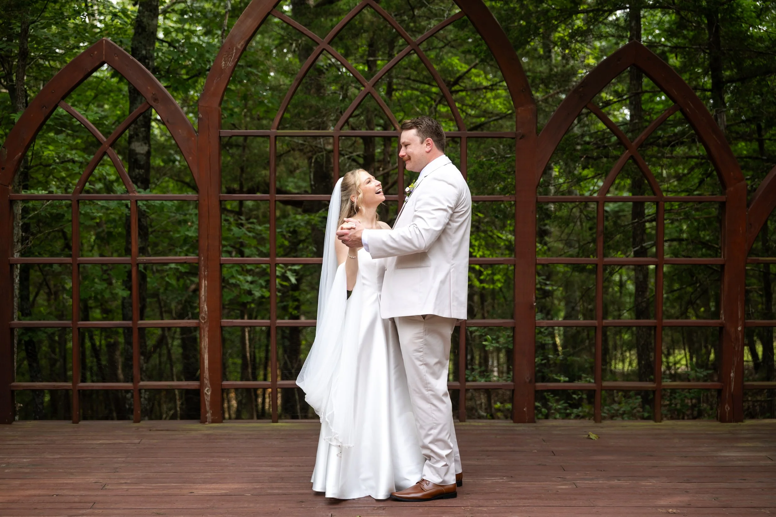 A bride and groom share a moment outdoors, standing on a wooden deck with trees in the background. The bride is wearing a white wedding dress and veil, and the groom is dressed in a white suit and brown shoes. They are smiling and looking at each other.