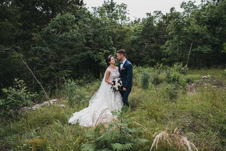 Bride and groom standing close together in a grassy, wooded outdoor setting, the bride in a white wedding dress holding a bouquet, and the groom in a dark blue suit, smiling and looking at each other.
