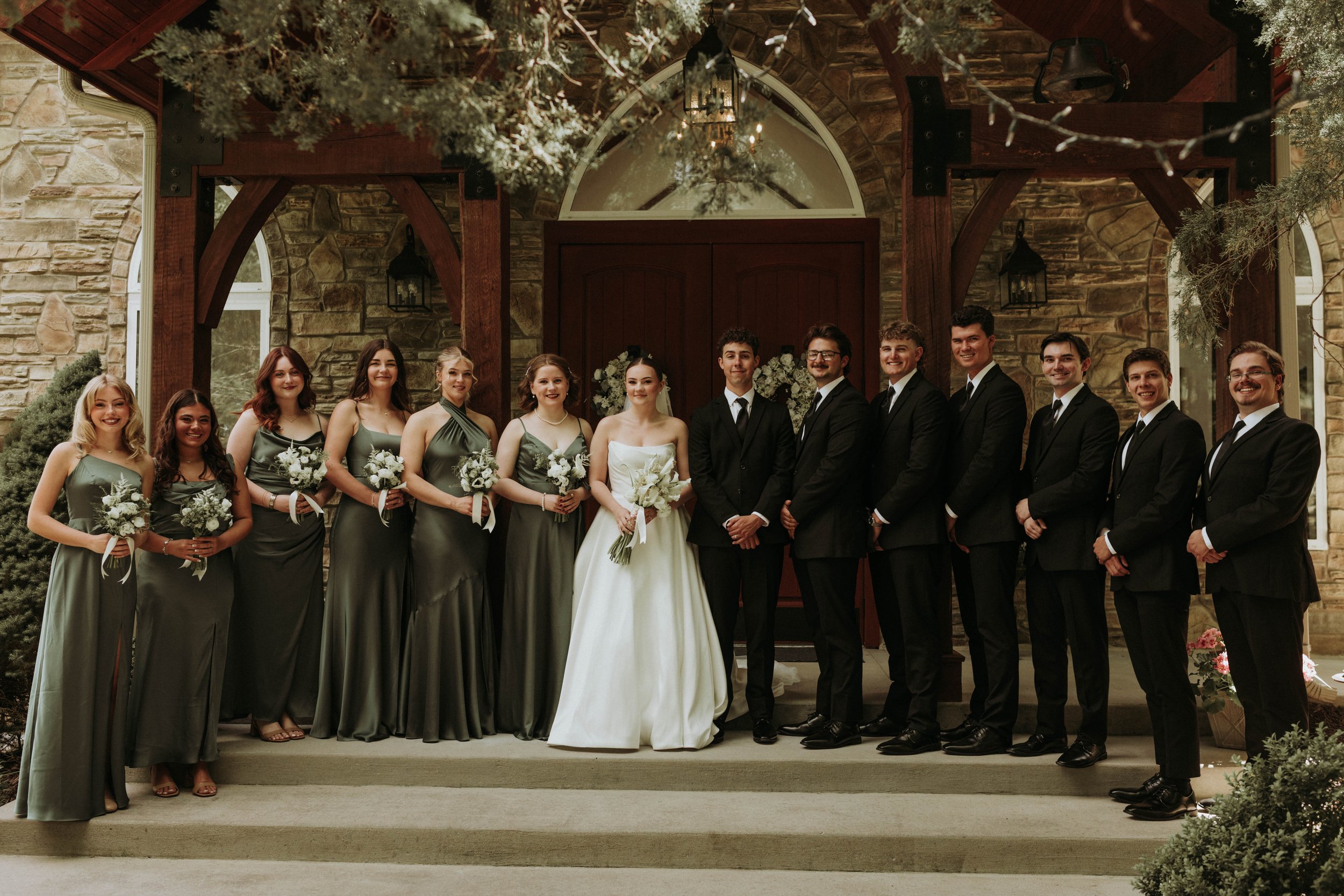 Group of people at a wedding, with women wearing dark gray dresses holding bouquets and men in black suits standing outdoors in front of a stone building with wooden beams and arched windows.