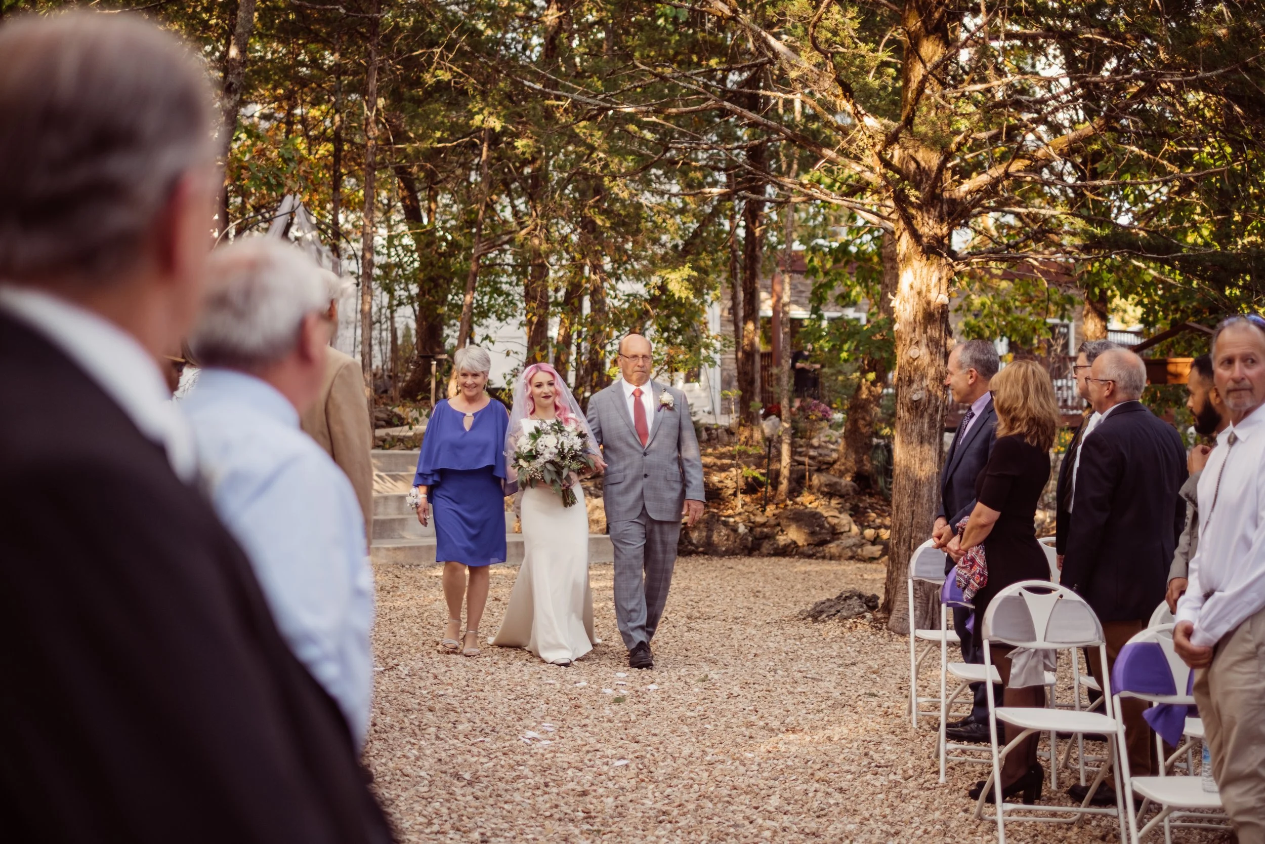 A bride walking down the outdoor aisle accompanied by her father during a wedding ceremony. Guests are seated on either side, and trees surround the scene in a natural setting.
