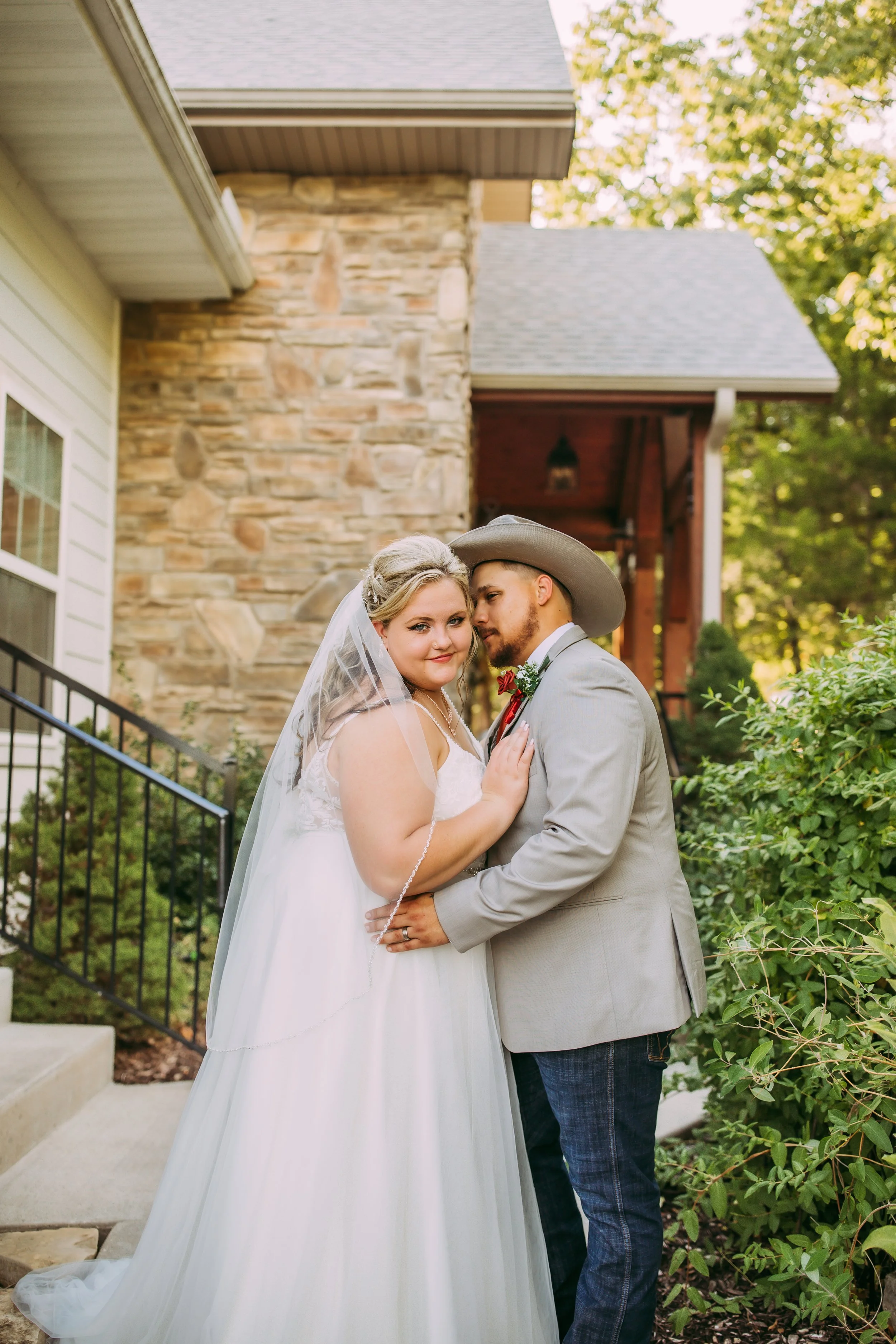 A bride and groom in wedding attire standing closely together outdoors in a garden, with a house featuring stone and siding walls in the background, surrounded by trees and greenery.