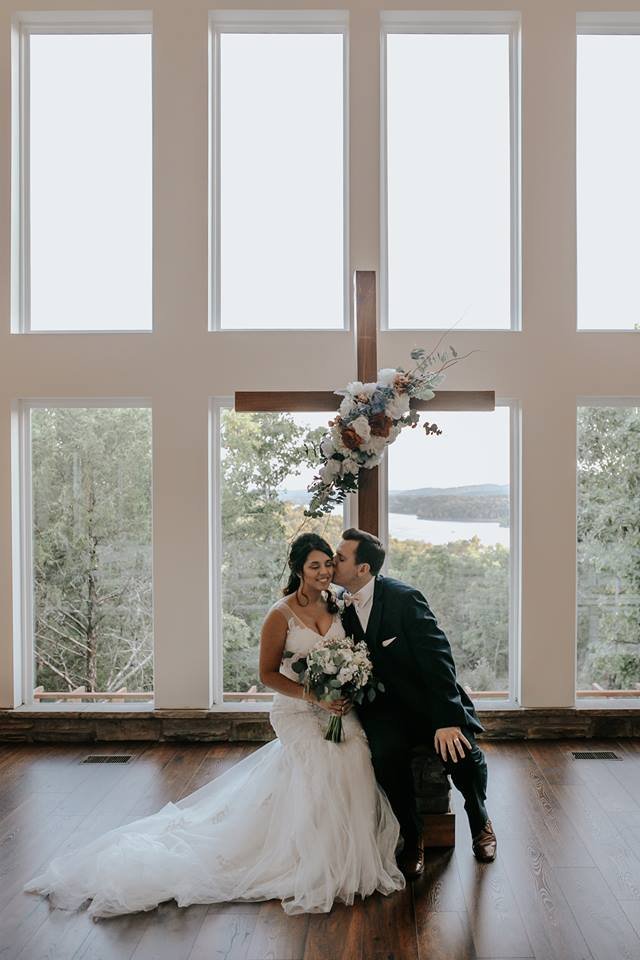 A bride and groom are inside a church, standing in front of a large cross decorated with flowers, with large windows and a view of trees and water outside. The bride is holding a bouquet of flowers, and the groom is sitting on a stool, giving her a k
