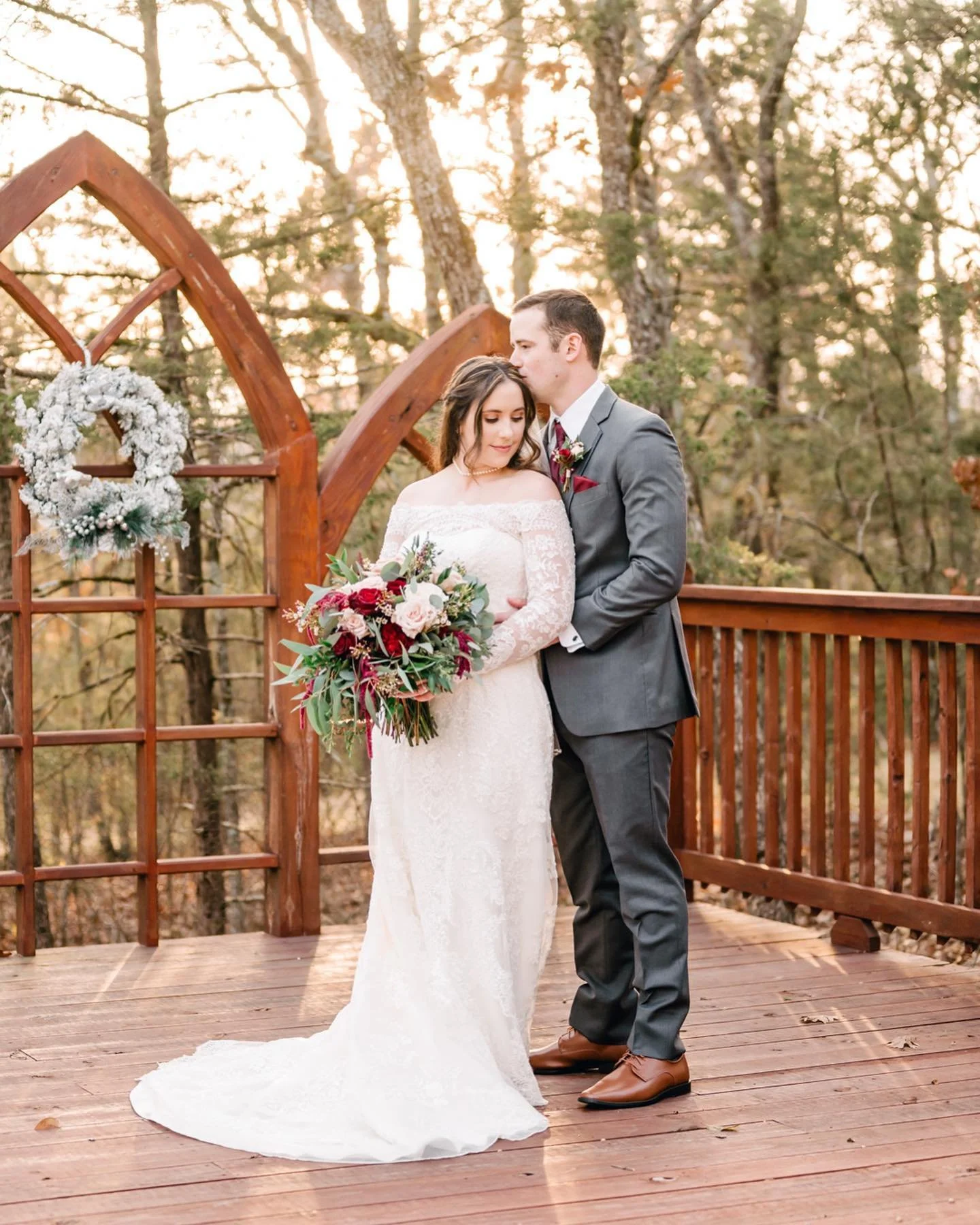A bride and groom standing together on a wooden deck outdoors during sunset, with trees in the background. The bride is holding a bouquet of flowers and wearing an off-the-shoulder white wedding dress, while the groom is dressed in a gray suit with a