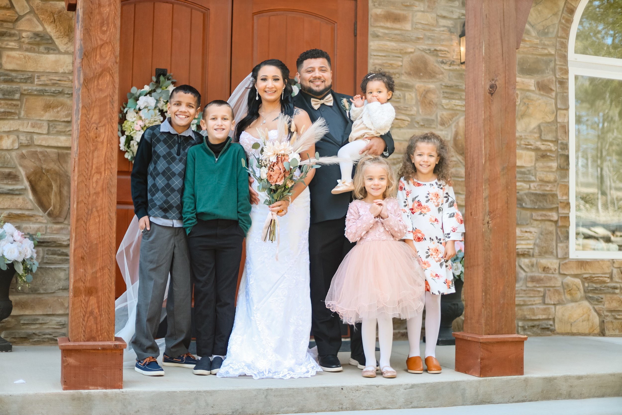 A wedding celebration with a group of six children and two adults, likely a bride and groom, standing on the steps outside a stone building with large windows. The bride is holding a bouquet and wearing a white wedding dress, while the man beside her