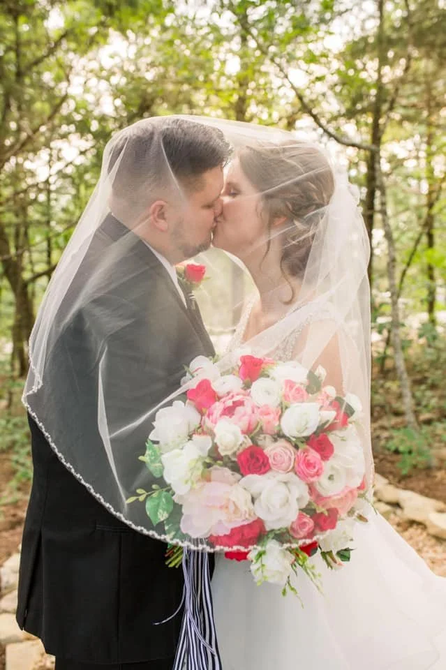 A newlywed couple sharing a kiss outdoors, with the bride holding a large bouquet of pink, white, and red roses, covered by a veil, in a wooded area.