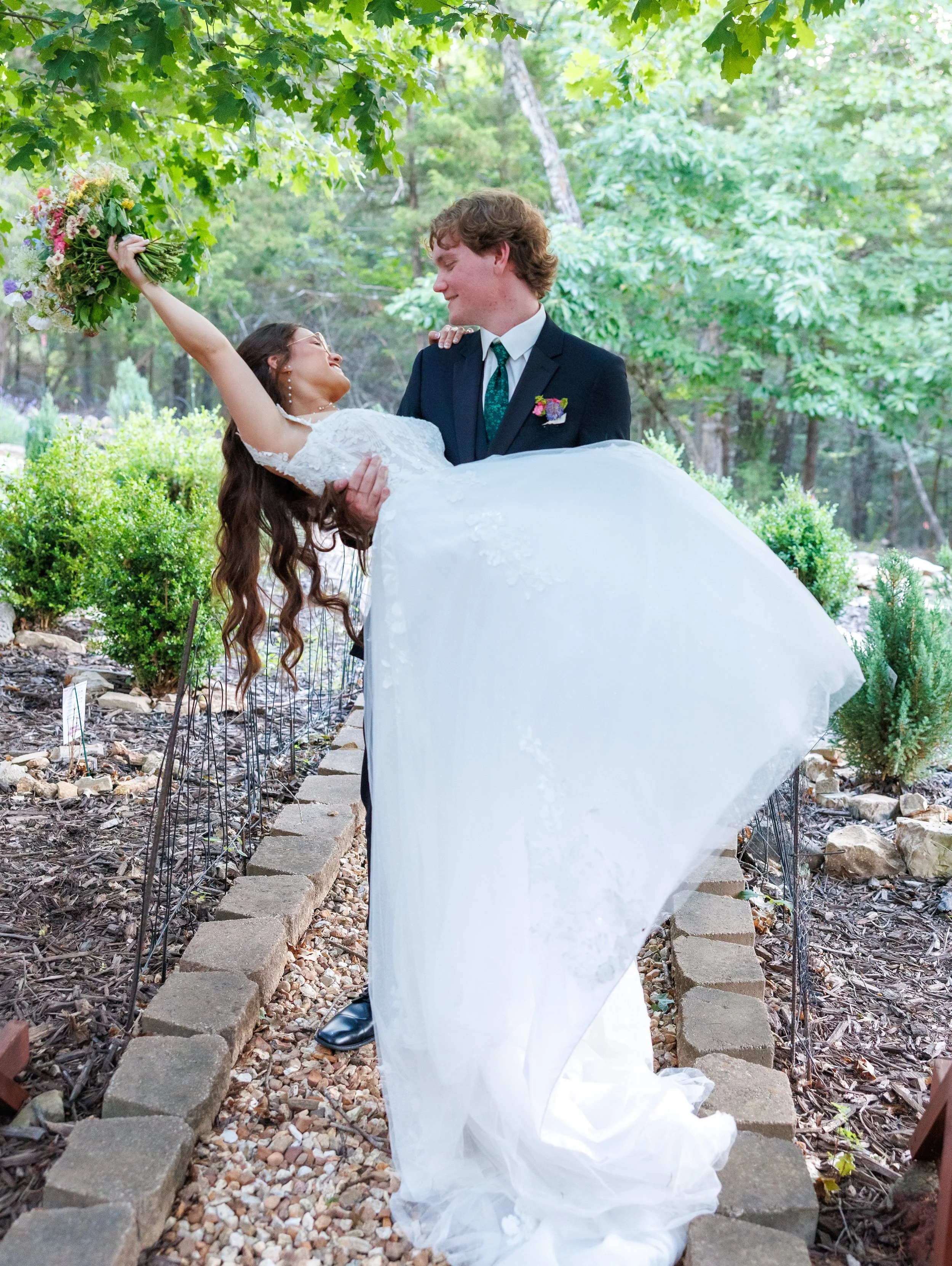 A bride and groom in wedding attire are outdoors, standing on a stone pathway amidst trees. The groom is holding the bride who is smiling and extending a bouquet of flowers upward, creating a joyful scene.